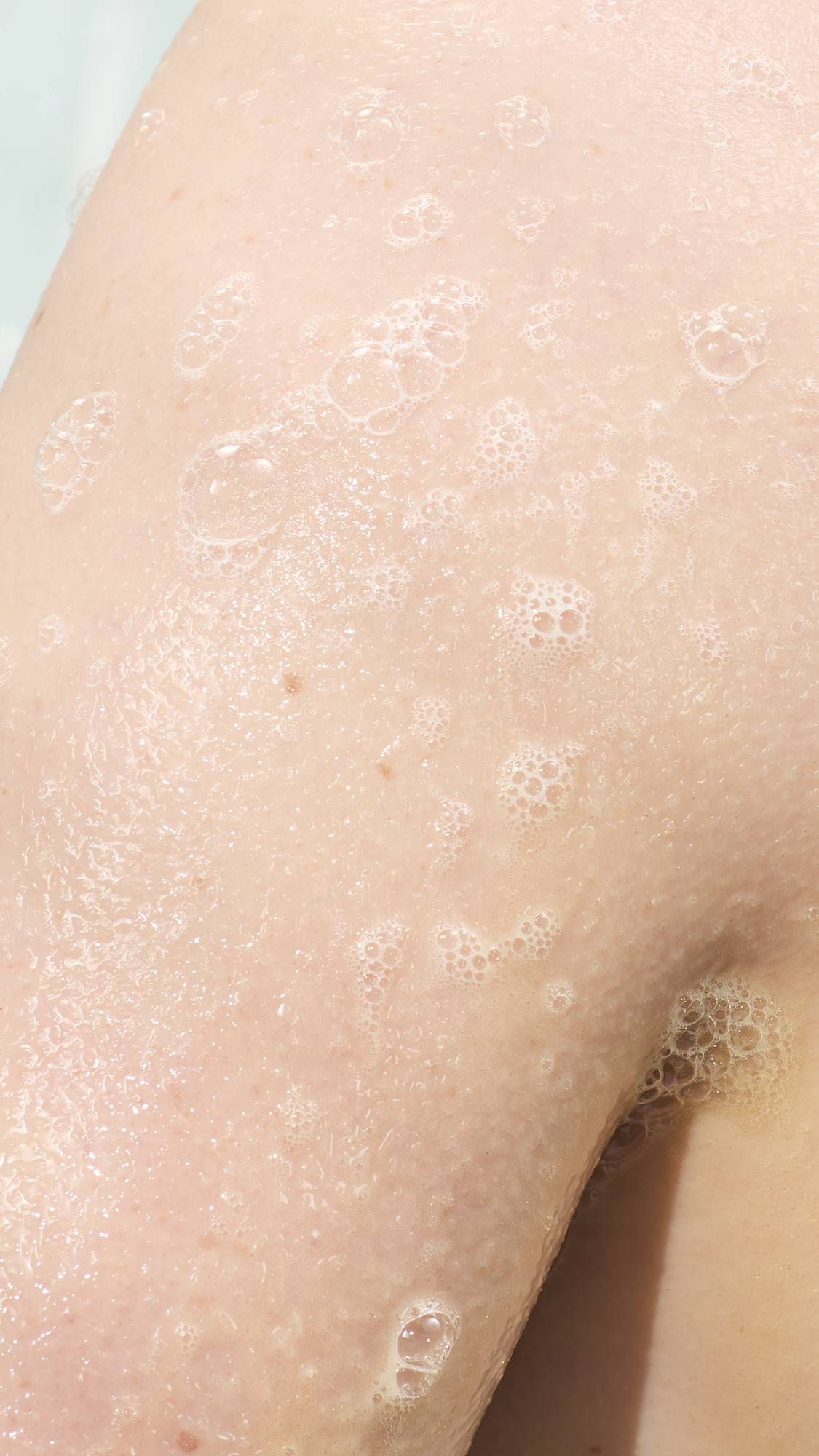 A close-up of a person's skin in the shower, covered in a thin layer of shimmering gold-tinted soap suds. Small, delicate white bubbles are scattered across the damp skin, creating a glistening, hydrated texture.