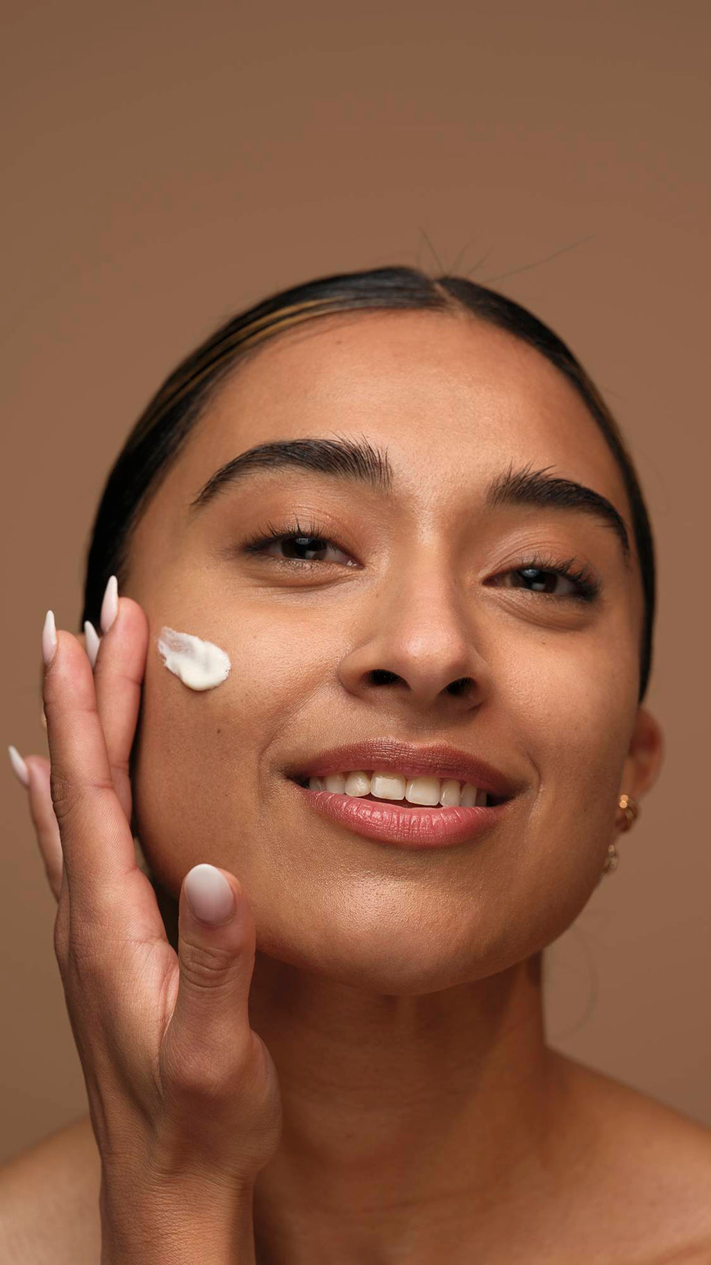 A close-up image of the model's smiling face as they start to apply the Gorgeous self-preserving moisturiser to one cheek. They are on a warm, earthy-brown background.