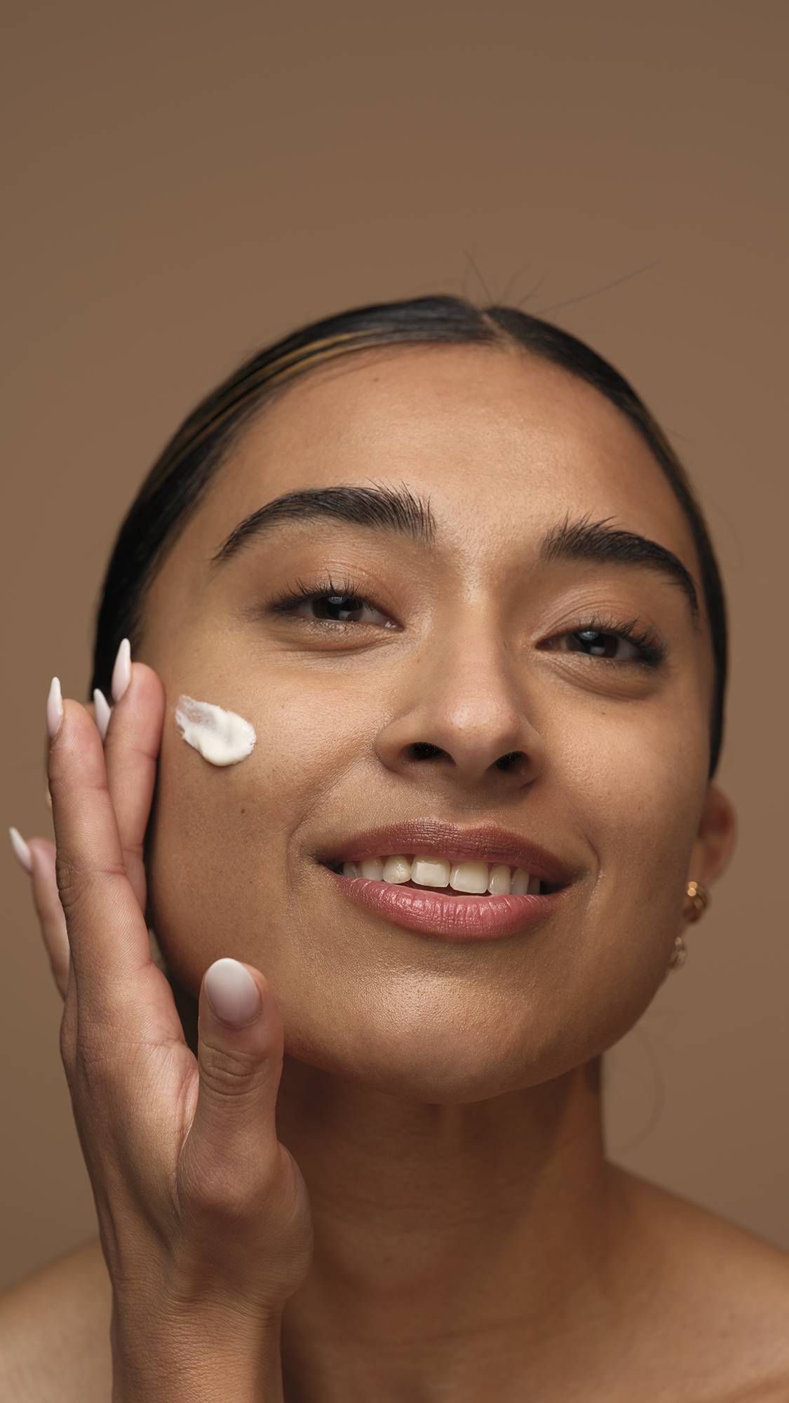 A close-up image of the model's smiling face as they start to apply the Gorgeous self-preserving moisturiser to one cheek. They are on a warm, earthy-brown background.