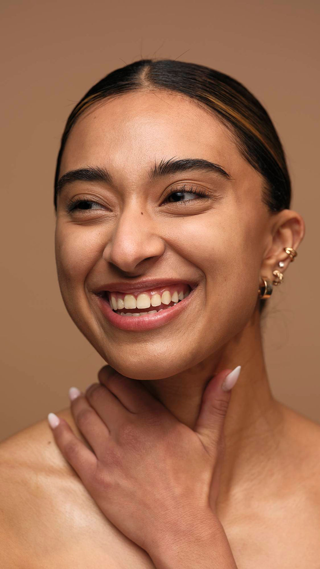 The model is on a warm, earthy-brown background with their hand across their chest and neck as they are smiling with glowing, freshly moisturised skin.