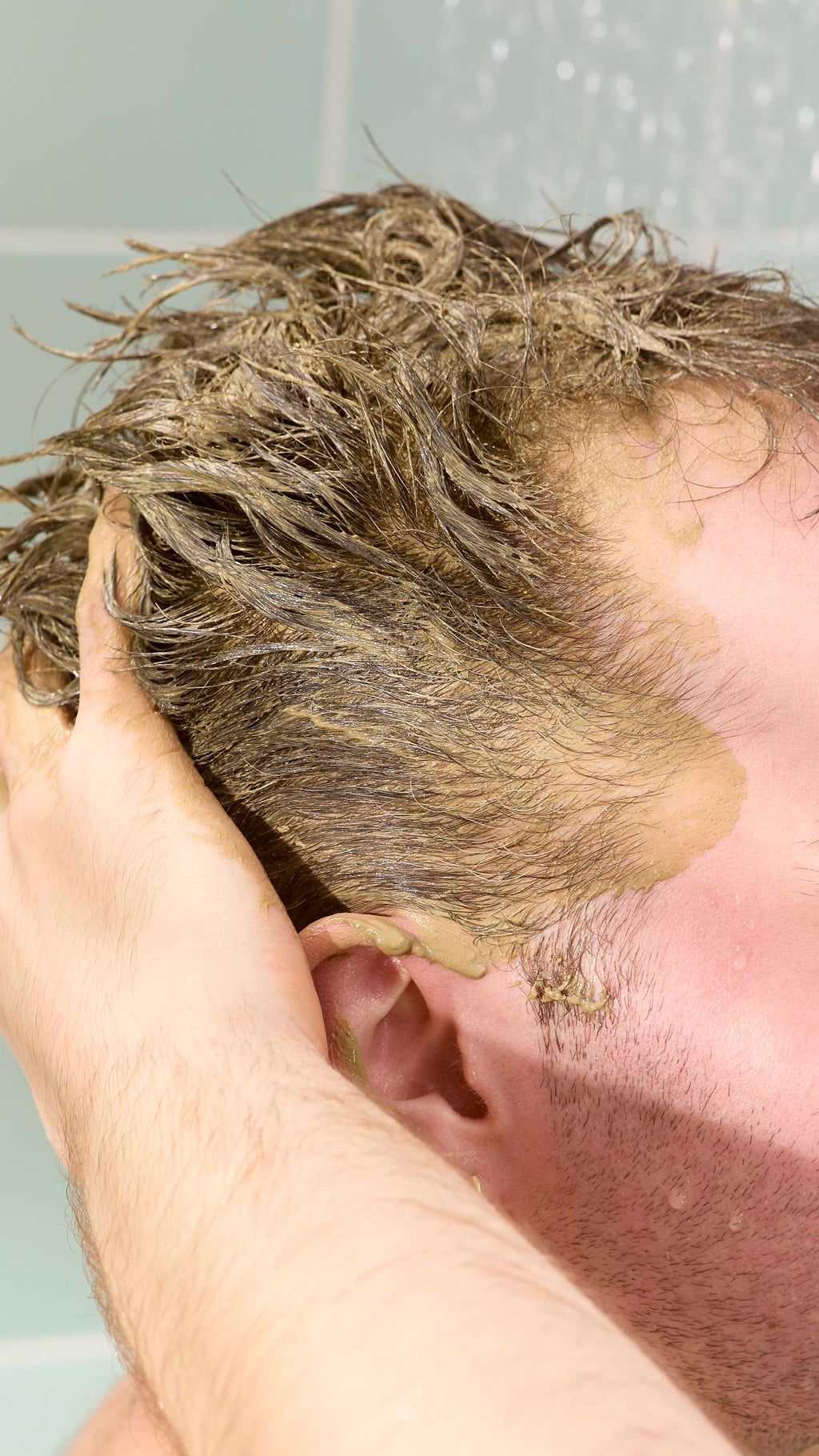 A close-up of a model applying a thick, olive-green "Green Shoots" hair food treatment to their short, dark blonde hair. Their hand is pressed against the side of their head, with the paste visible on their temple, ear, and through the hair strands.