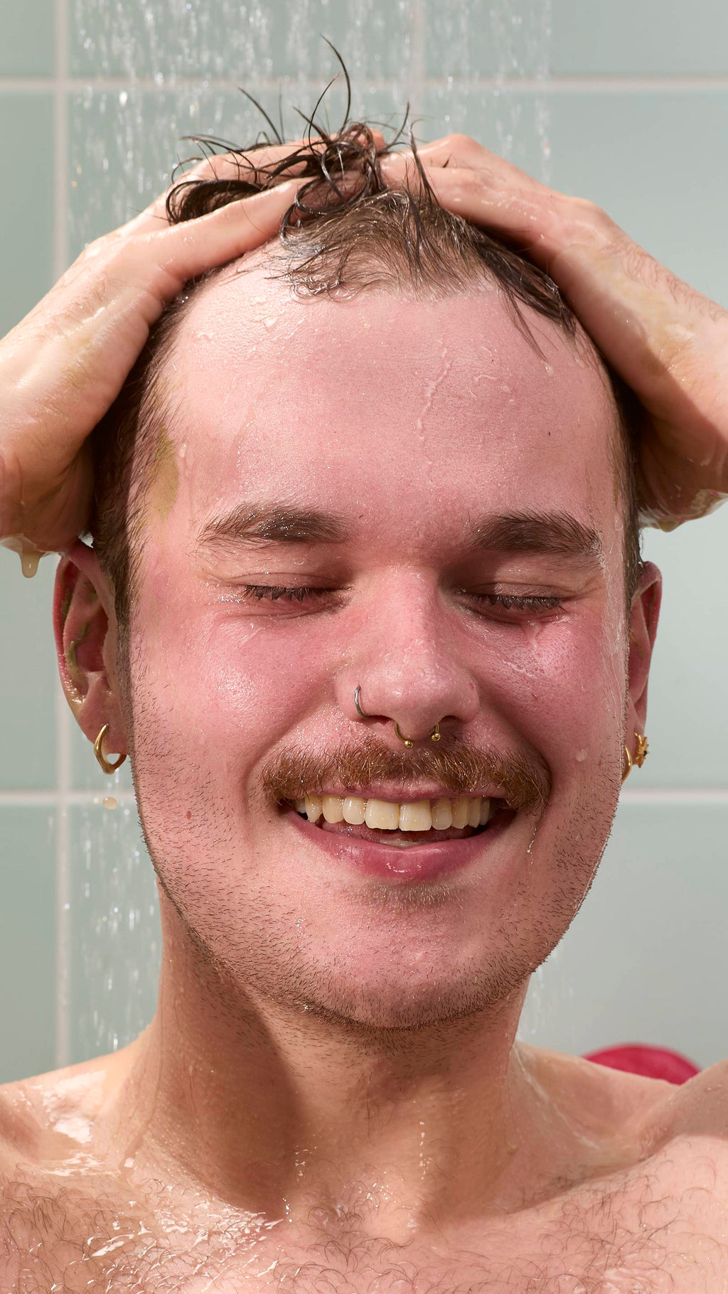 A close-up of a model with a moustache and nose piercings smiling with eyes closed under a shower. They are rinsing off an olive-green "Green Shoots" hair food treatment. Water droplets fall against a background of light blue wall tiles.