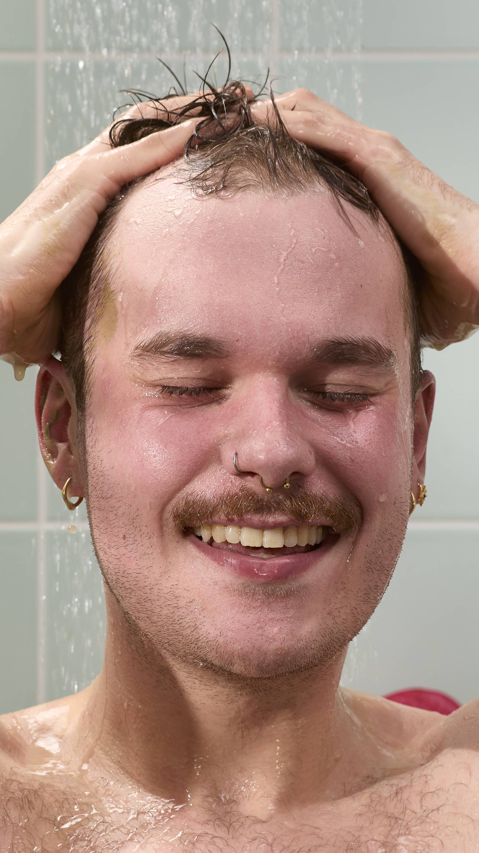 A close-up of a model with a moustache and nose piercings smiling with eyes closed under a shower. They are rinsing off an olive-green "Green Shoots" hair food treatment. Water droplets fall against a background of light blue wall tiles.