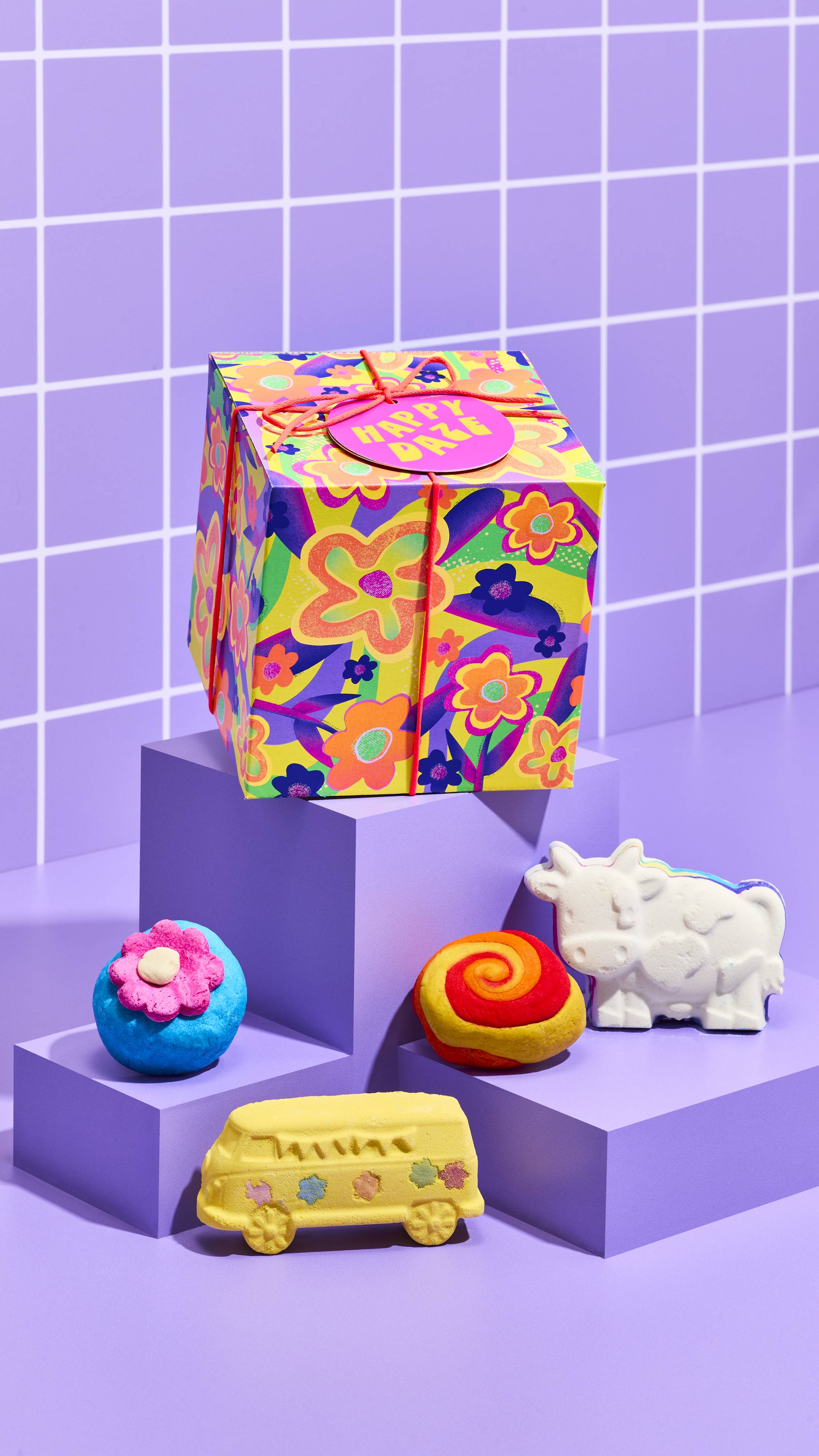 The image shows the colourful neon flower-designed Happy Daze gift box, sitting on a cubed platform with the four included bath products sitting on lower platforms on a pastel-purple tiled background.