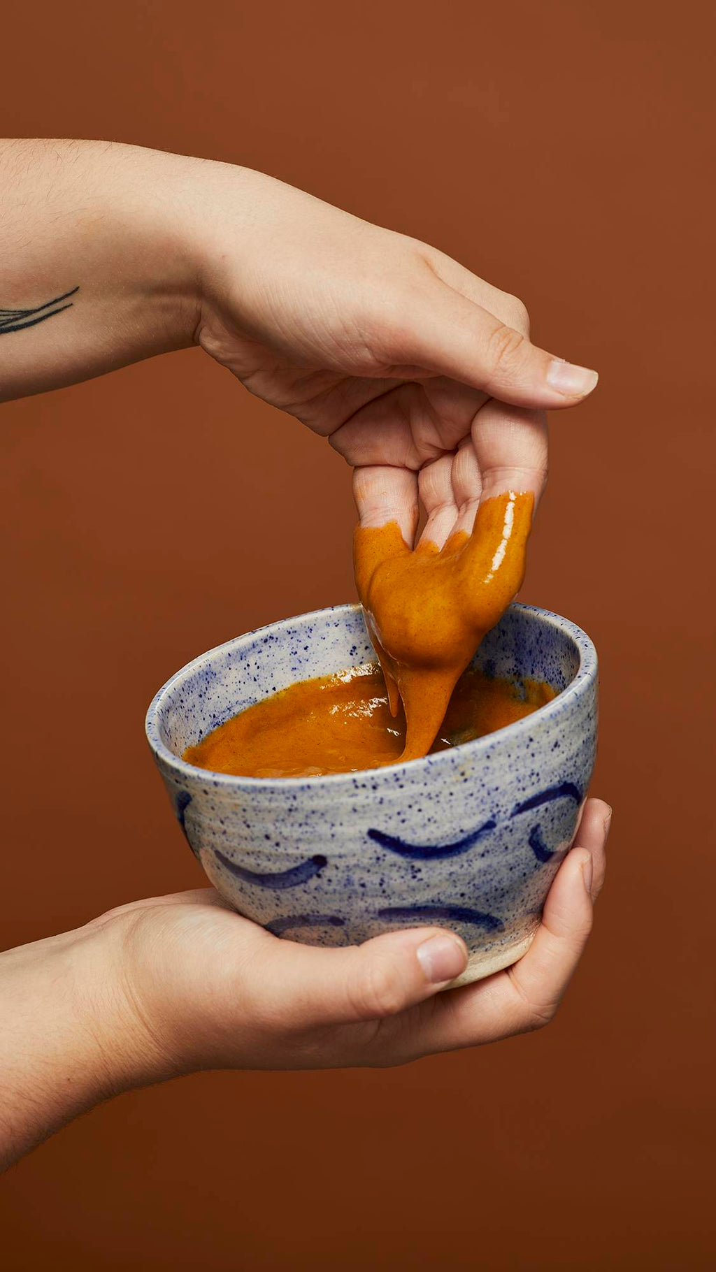 Image shows the model scooping the Hennaed hot oil treatment out of a blue ceramic bowl in front of an earthy background.