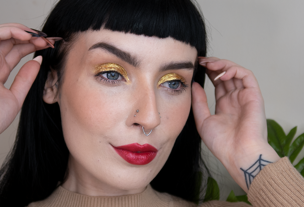 A person with dark hair pouts, their eyelids covered in shimmery gold Fantasy eyeliner and their lips in bright red lipstick.