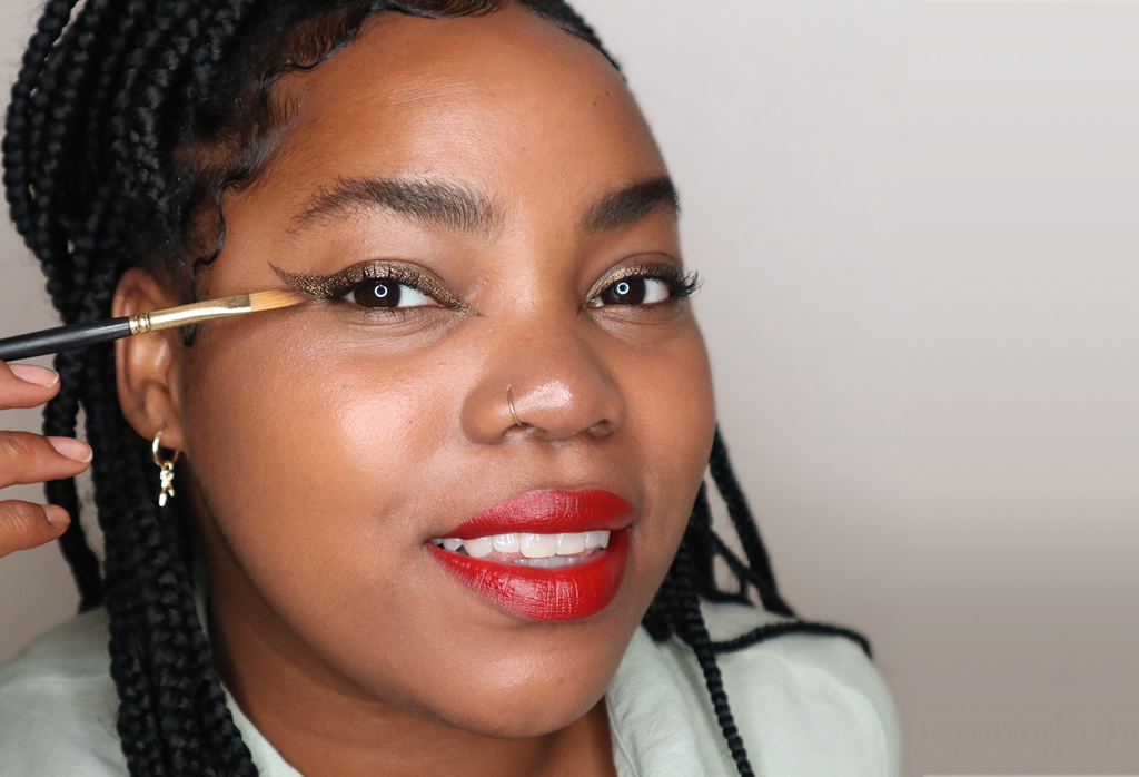 A person with braids smiles, as they apply shimmery bronze eyeliner Grounded with a Wing It Brush.
