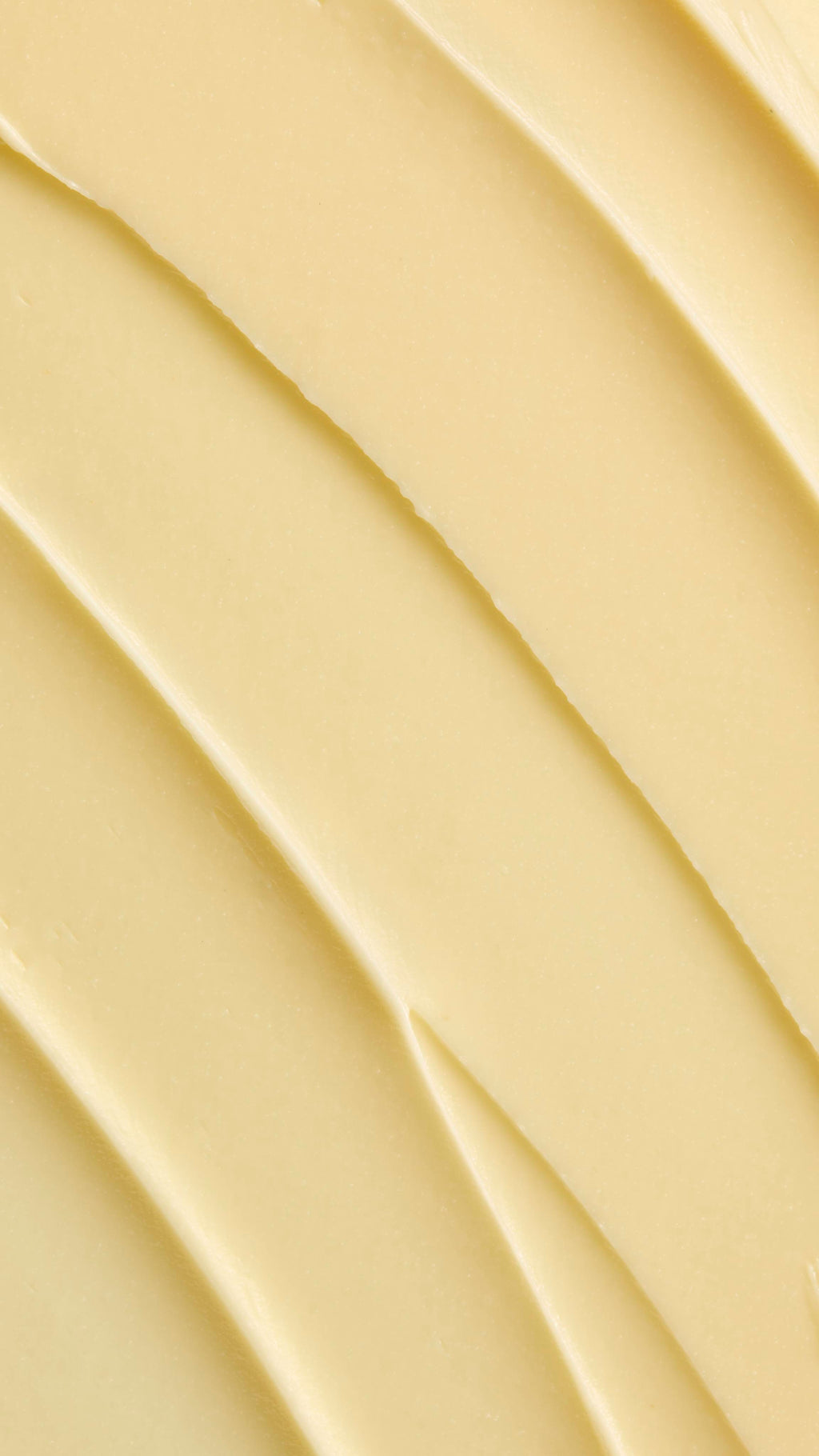 A super close-up image showing diagonal, overlapping smears of the thick, creamy, pale yellow Lush "Honey, Beeswax And Almond Oil" facial cleanser. The smooth product has a matte finish and subtle ridged textures.