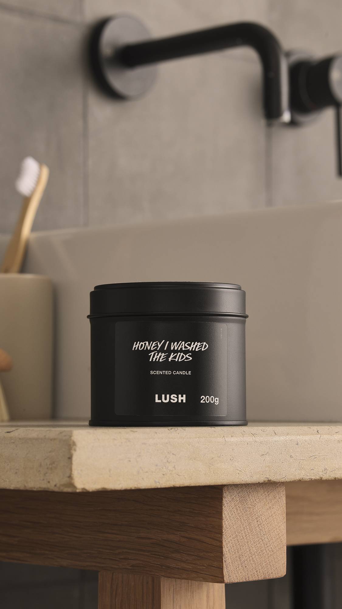The image shows a blurred, grey background with a black tap and bamboo toothbrush. In the foreground, the Honey I Washed The Kids black tin candle with the lid sits atop a wooden, stone-topped counter.