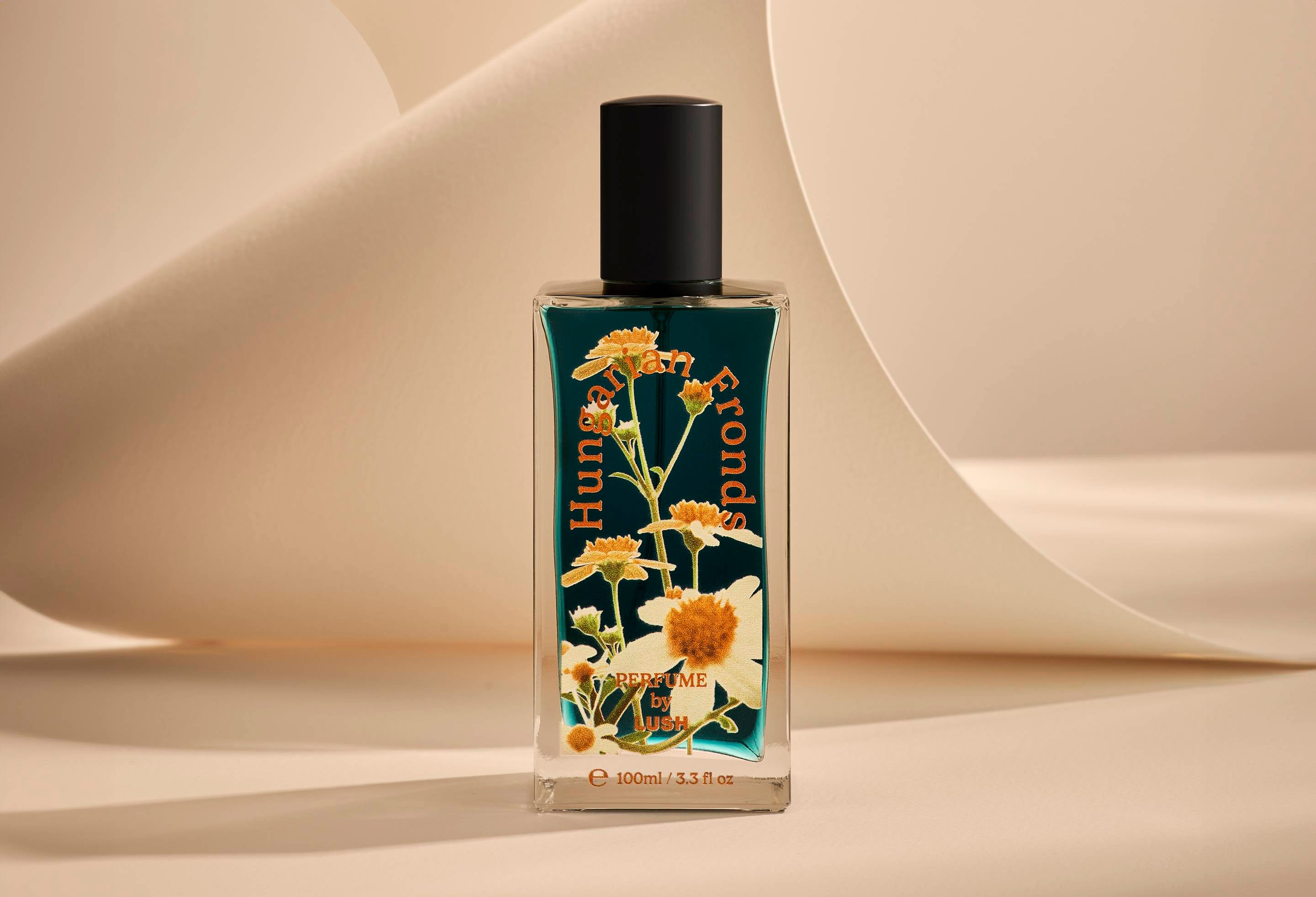 The image shows the Hungarian Fronds glass perfume bottle. It is sitting on a soft, beige background with curved, cream paper creating soft, gliding shapes behind the product.