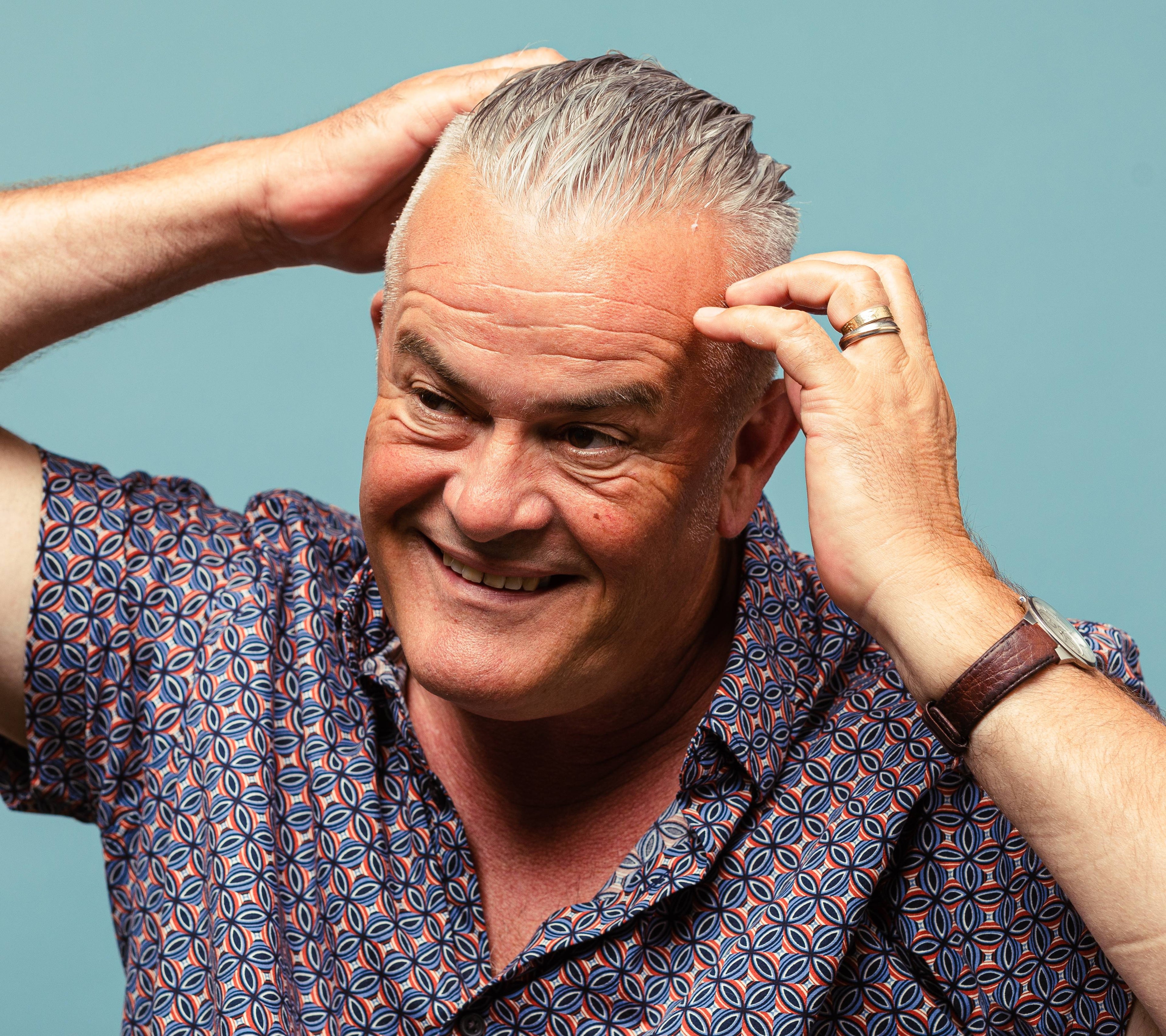 A person with short, grey hair and a jazzy shirt runs Infra Wig, a soft hair styling gel through the top section of their hair.