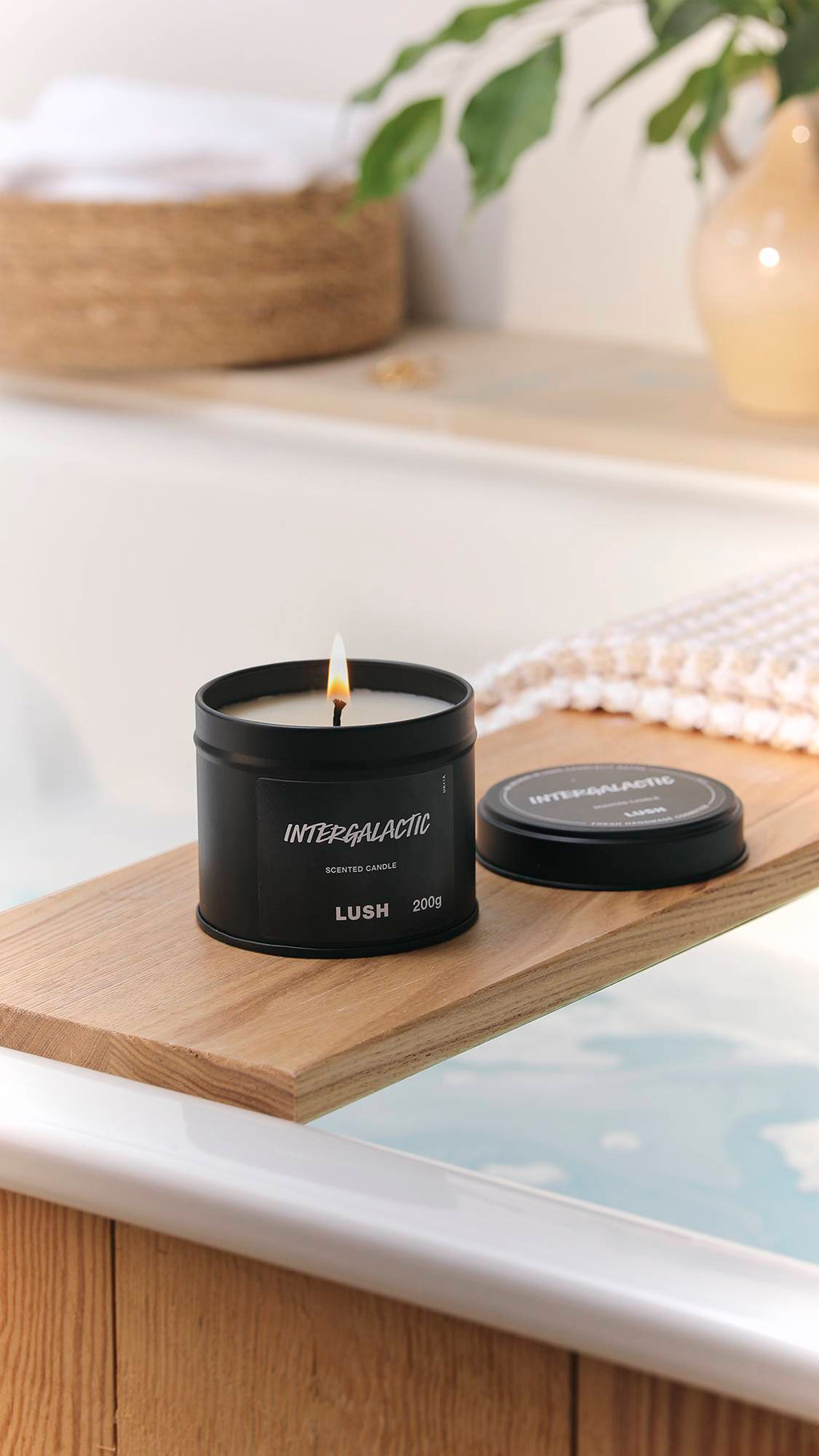 The image shows a bright, blurred bathroom scene with an out-of-focus wicker basket and plant. The black tin Intergalactic candle has a lit wick and is sitting on a wooden bath caddy laid over a filled bath.
