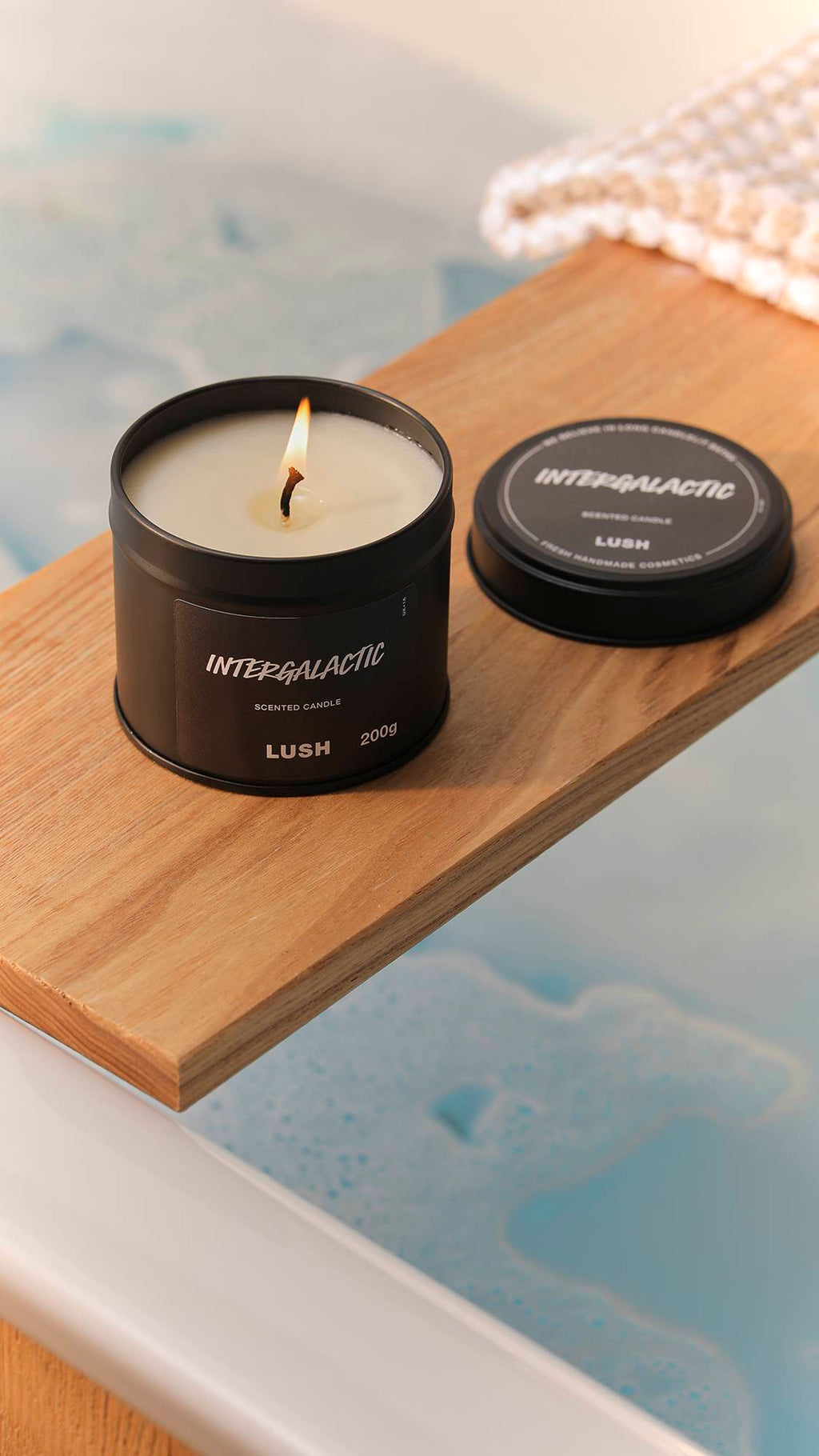 The image shows a bright, blurred bathroom scene. The black tin Intergalactic candle has a lit wick and is sitting on a wooden bath caddy laid over a filled bath.