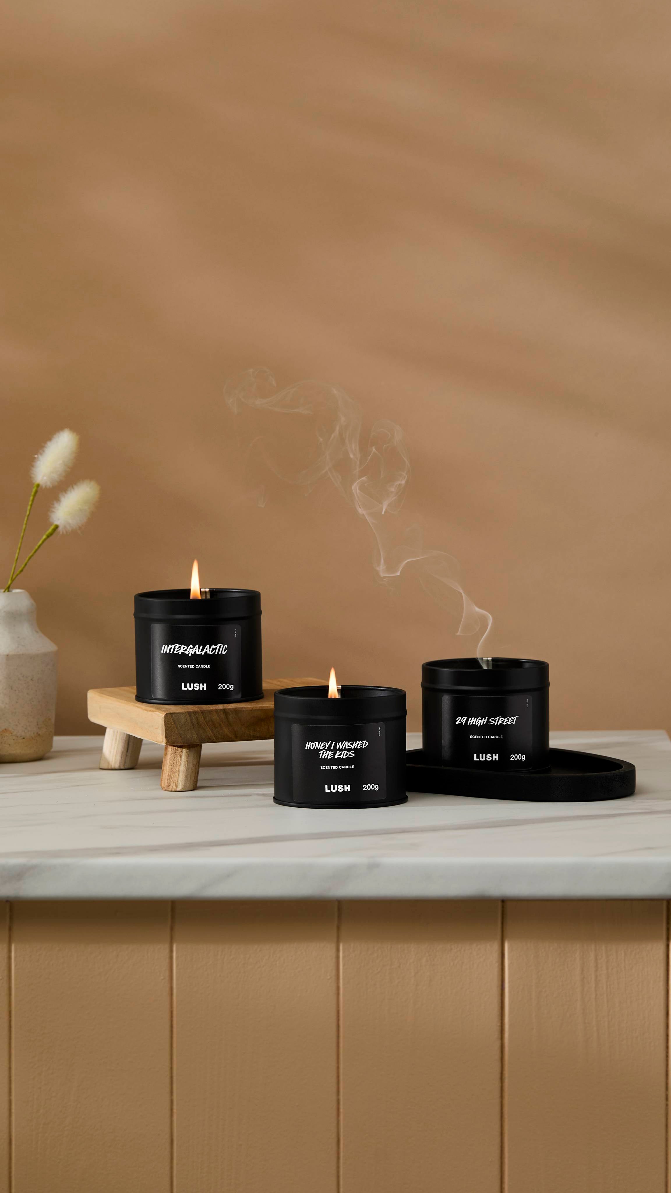 The image shows a warm beige background with a ceramic vase of Hare's Tail Grass. Three back tin candles are sitting with their wicks lit on a marble-topped counter with one having just been blown out with smoke rising,