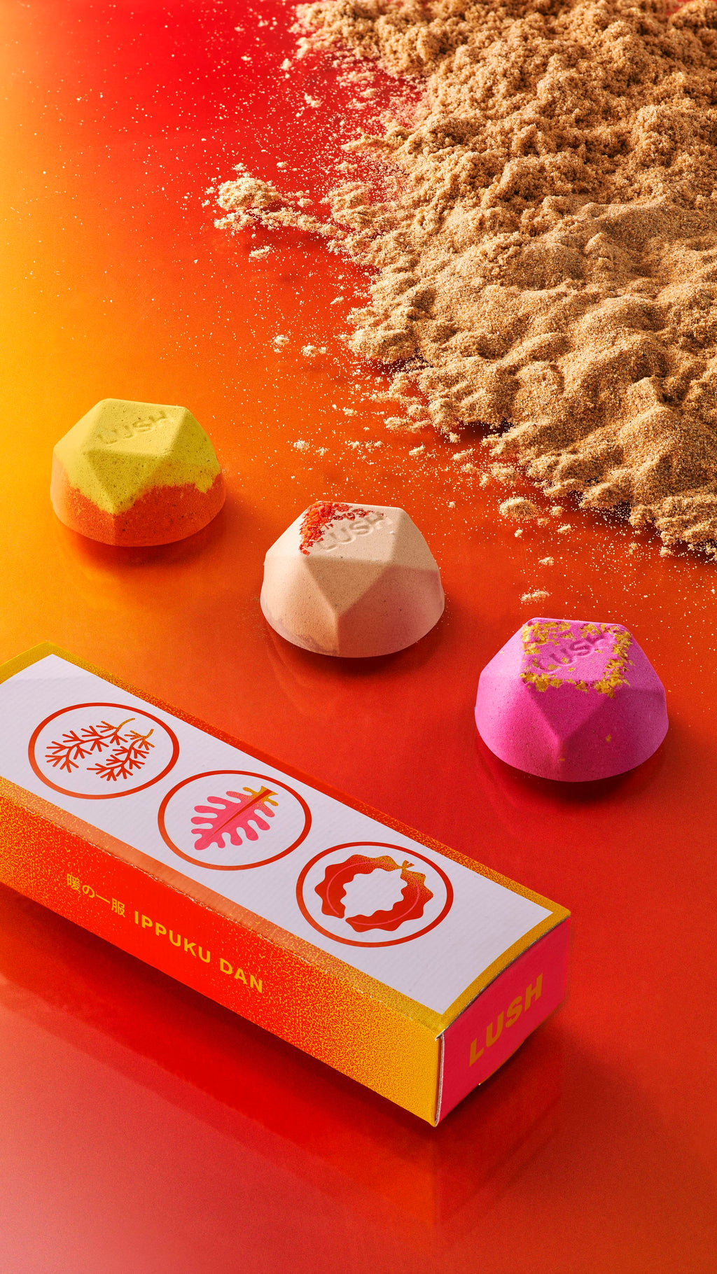 Three ippuku warming bath bombs lay in a line with sand on one side, the box on another on top of an orange-yellow gradient.