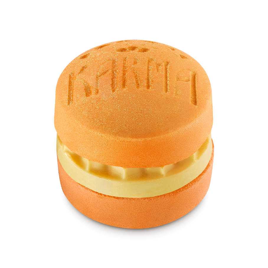 Karma. An orange, circular bath bomb shaped like a macaron with a creamy yellow middle layer of solid oil. The word 'KARMA' is embossed on the top surface, which is dusted with a fine gold shimmer.