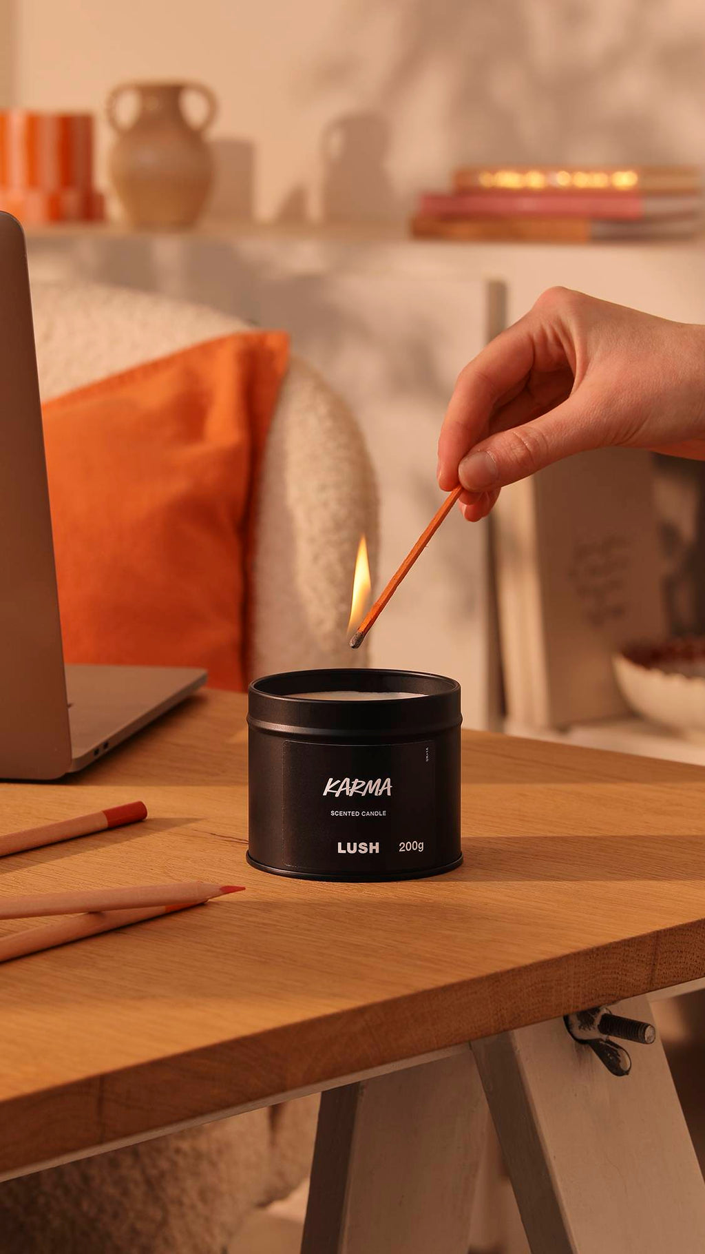 The image shows the black tin Karma candle sitting on a wooden desk as a model's hand reaches in to light the wick. The background is a warm, afternoon-sun lounge area with orange decor of books, pottery and pillows.