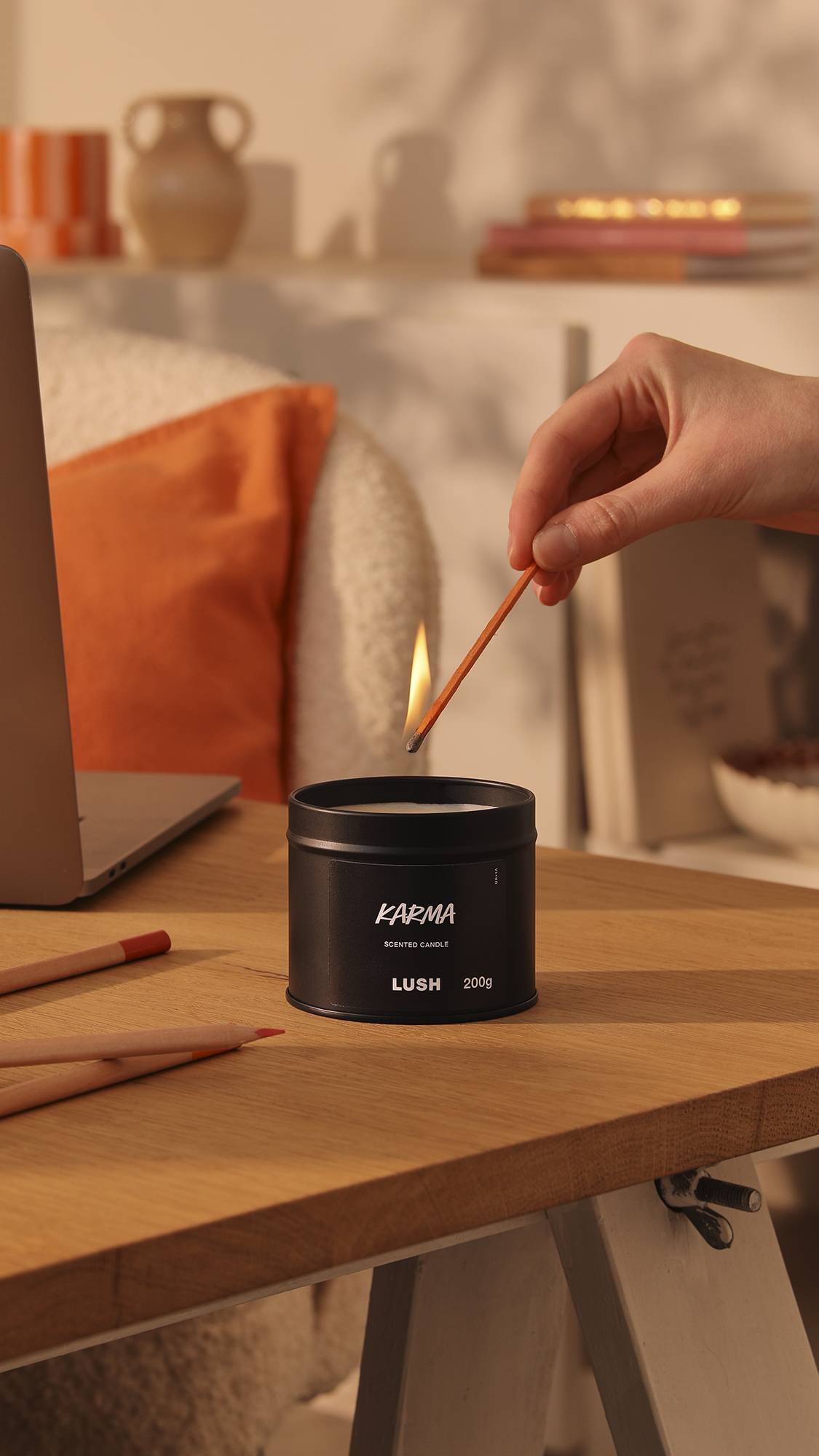 The image shows the black tin Karma candle sitting on a wooden desk as a model's hand reaches in to light the wick. The background is a warm, afternoon-sun lounge area with orange decor of books, pottery and pillows.