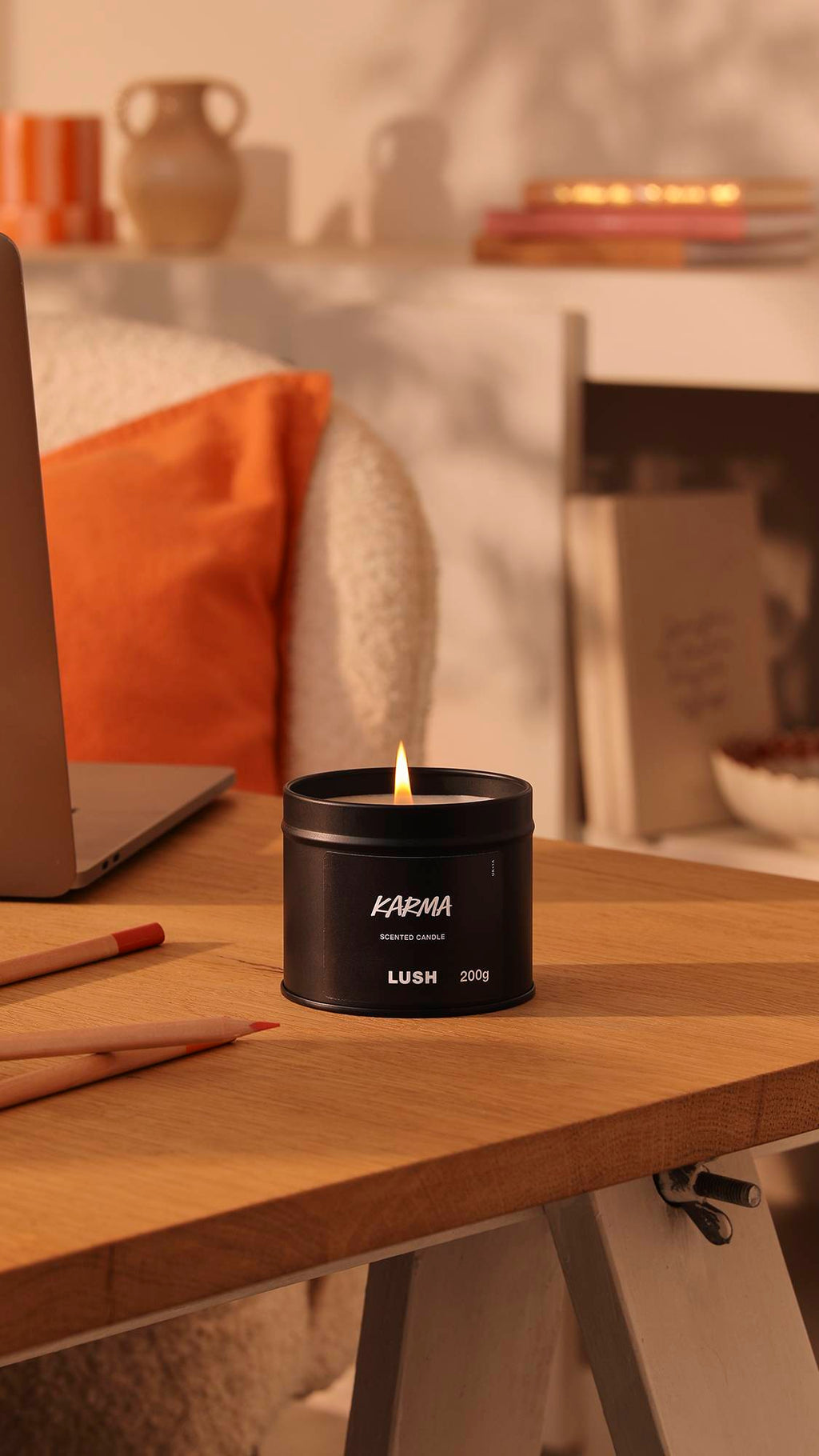 The image shows the black tin Karma candle with a lit flame sitting on a wooden desk. The background is a warm, afternoon-sun lounge area with orange decor of books, pottery and pillows.