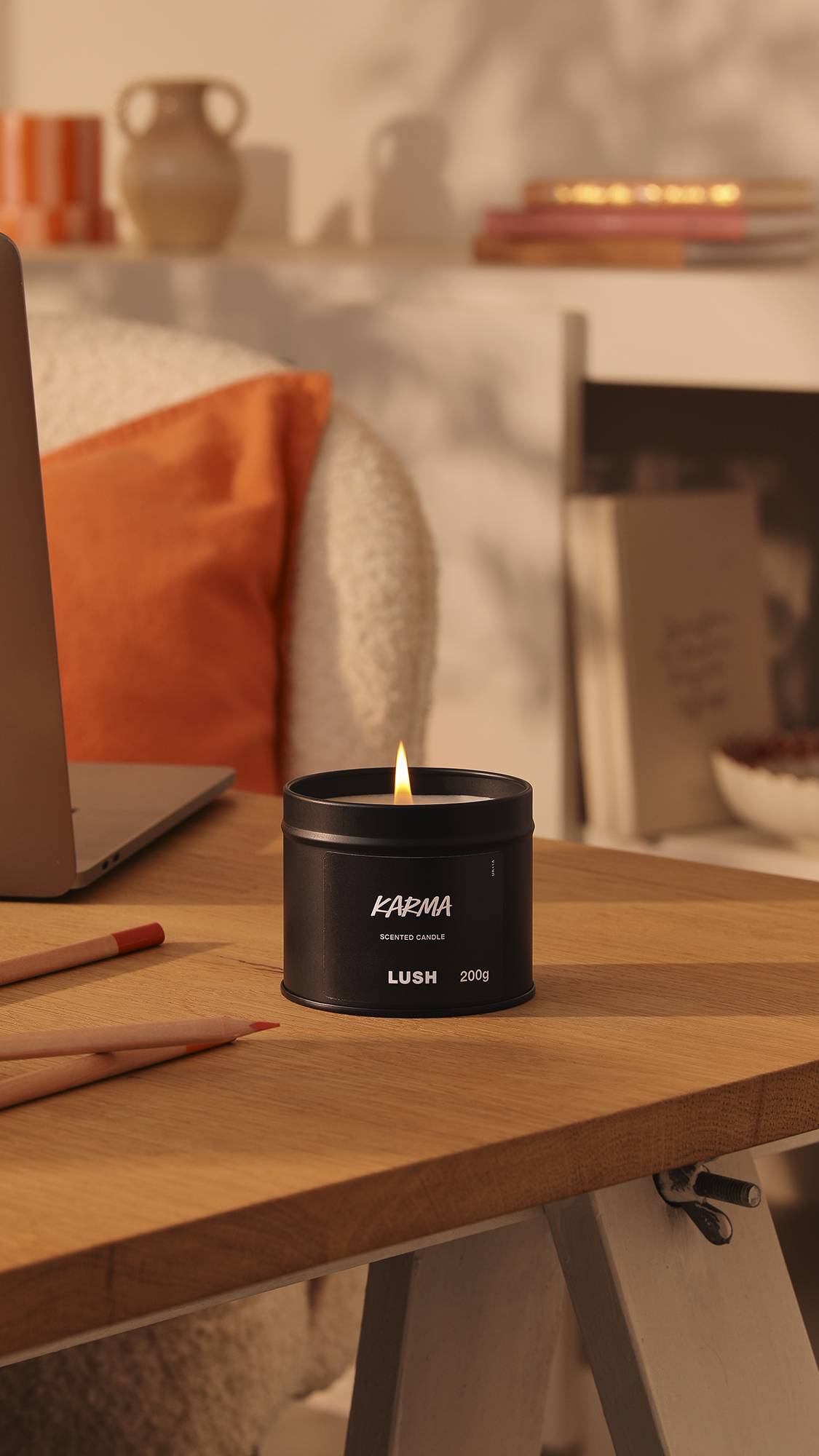 The image shows the black tin Karma candle with a lit flame sitting on a wooden desk. The background is a warm, afternoon-sun lounge area with orange decor of books, pottery and pillows.