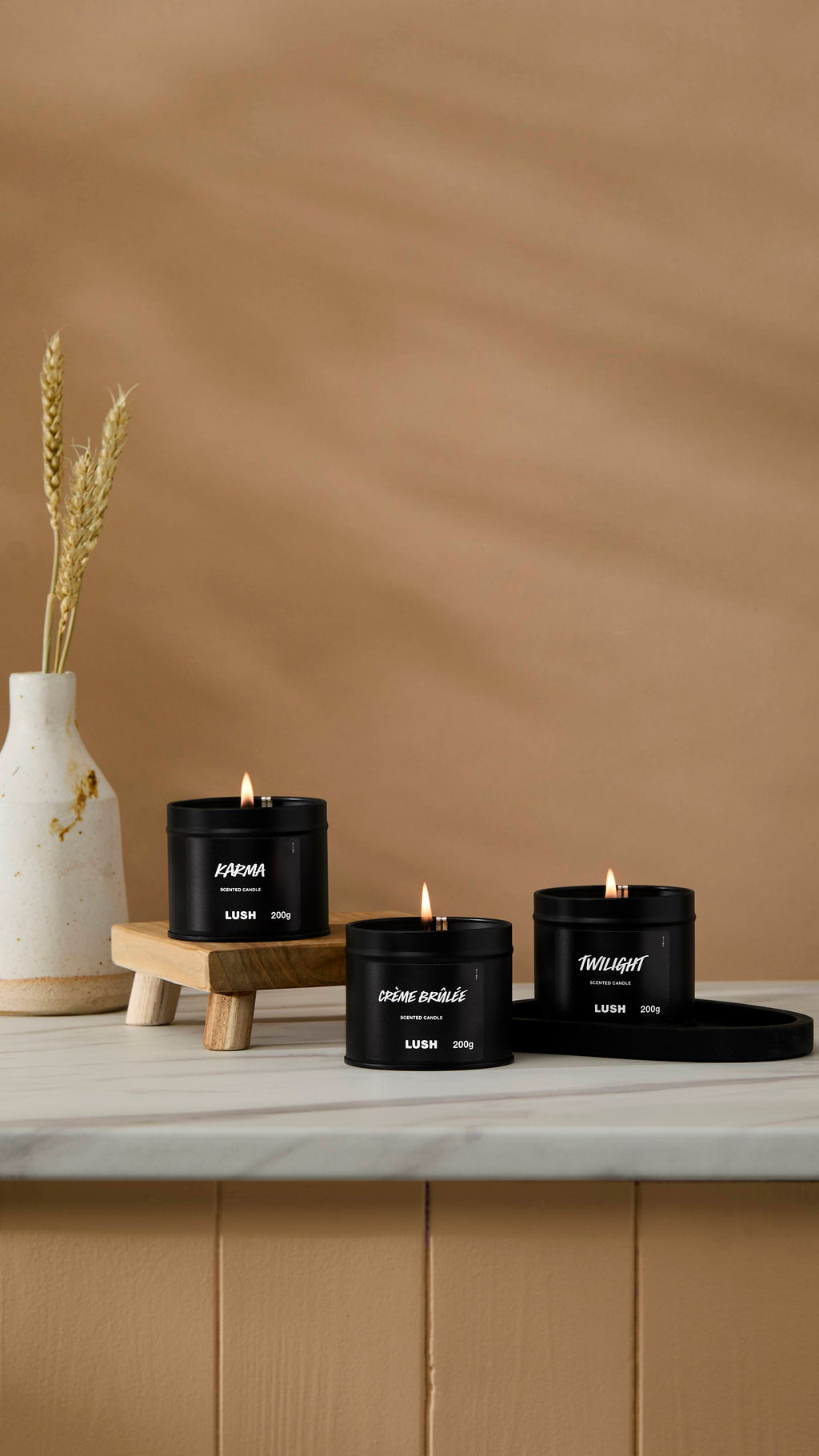 The image shows a warm beige background with a ceramic vase of wheat. Three back tin candles are sitting with their wicks lit on a marble-topped counter.