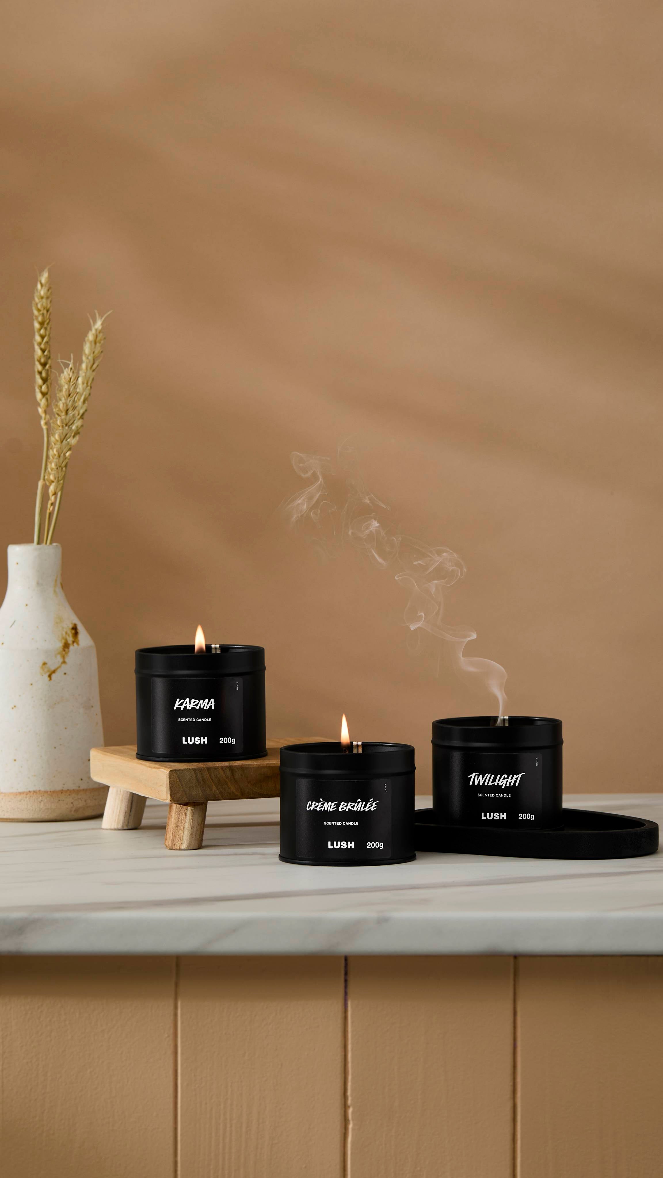 The image shows a warm beige background with a ceramic vase of wheat. Three back tin candles are sitting with their wicks lit on a marble-topped counter.