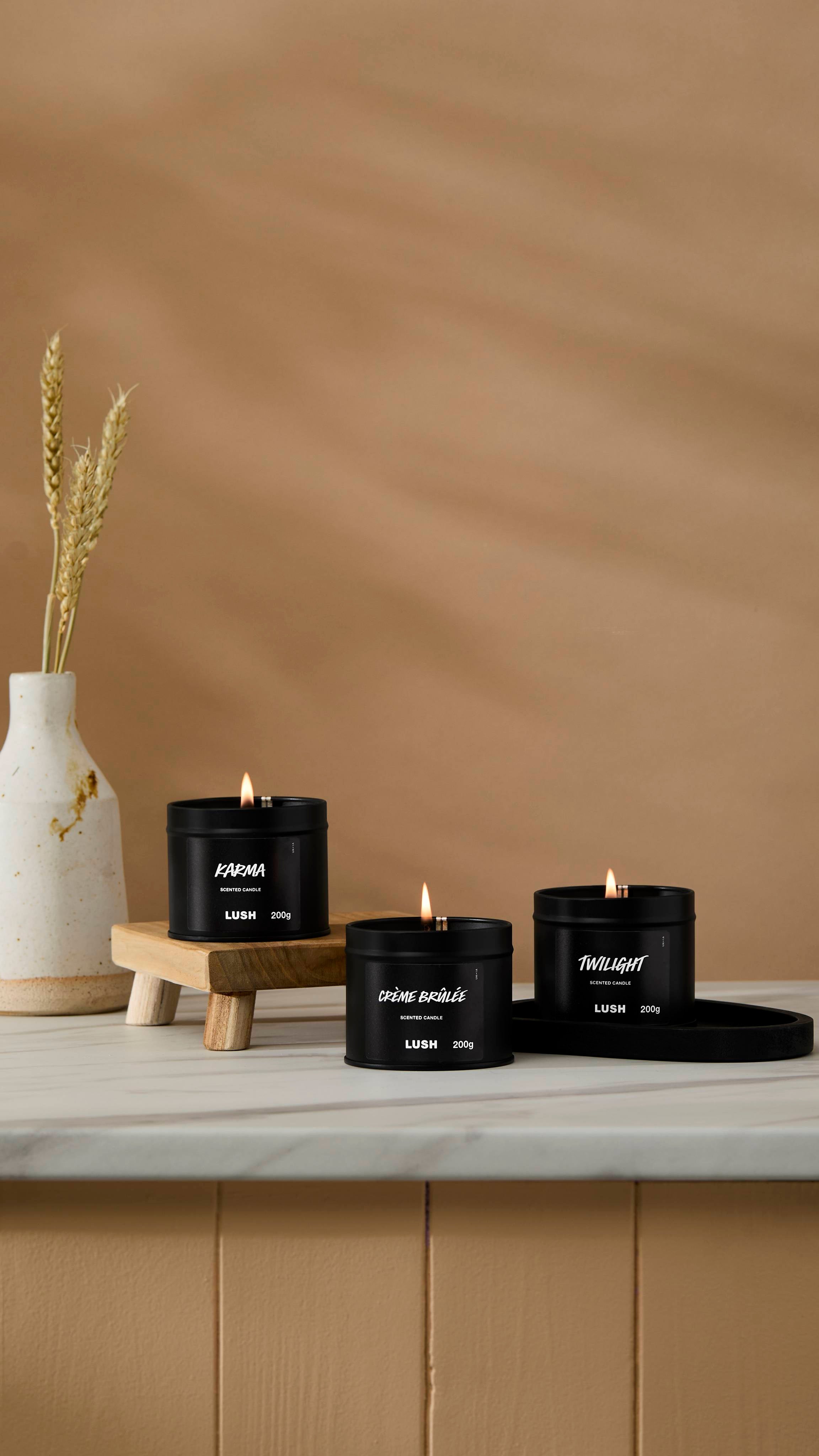 The image shows a warm beige background with a ceramic vase of wheat. Three back tin candles are sitting with their wicks lit on a marble-topped counter.