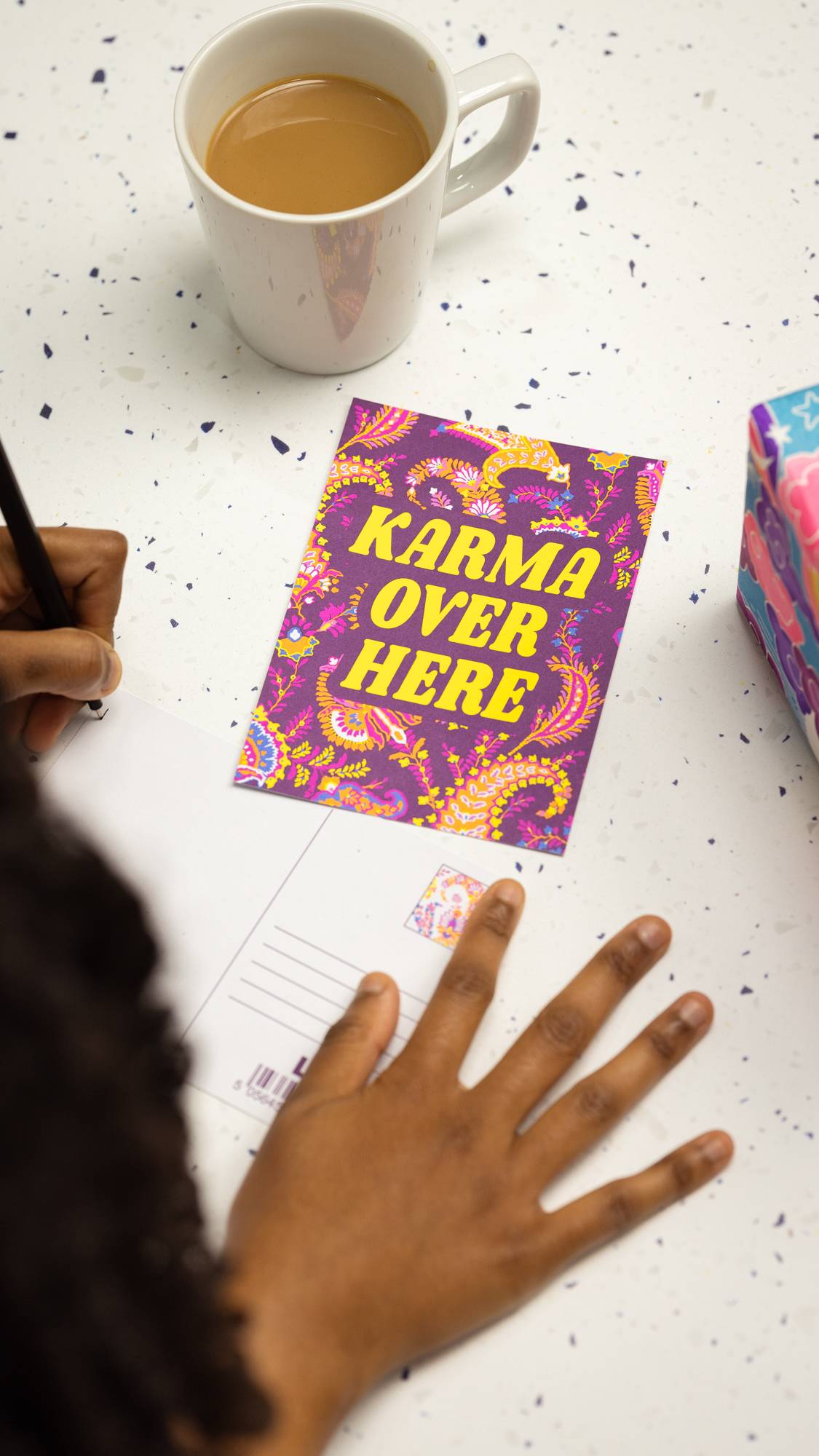 Image shows the Karma Over Here postcard laid flat on a table as the model writes on another postcard.