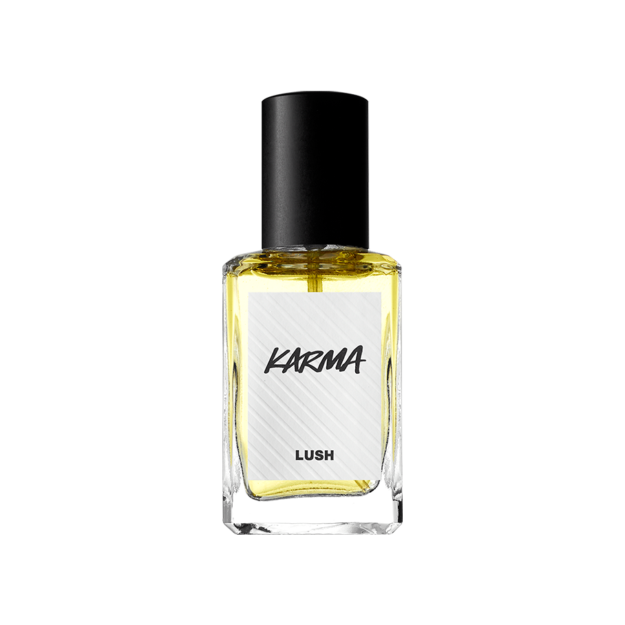 A rectangular glass bottle of Lush 'Karma' perfume with a black cap, filled with light yellow liquid, stands against a solid black background. The white textured label features the name "KARMA" in stylised black lettering.