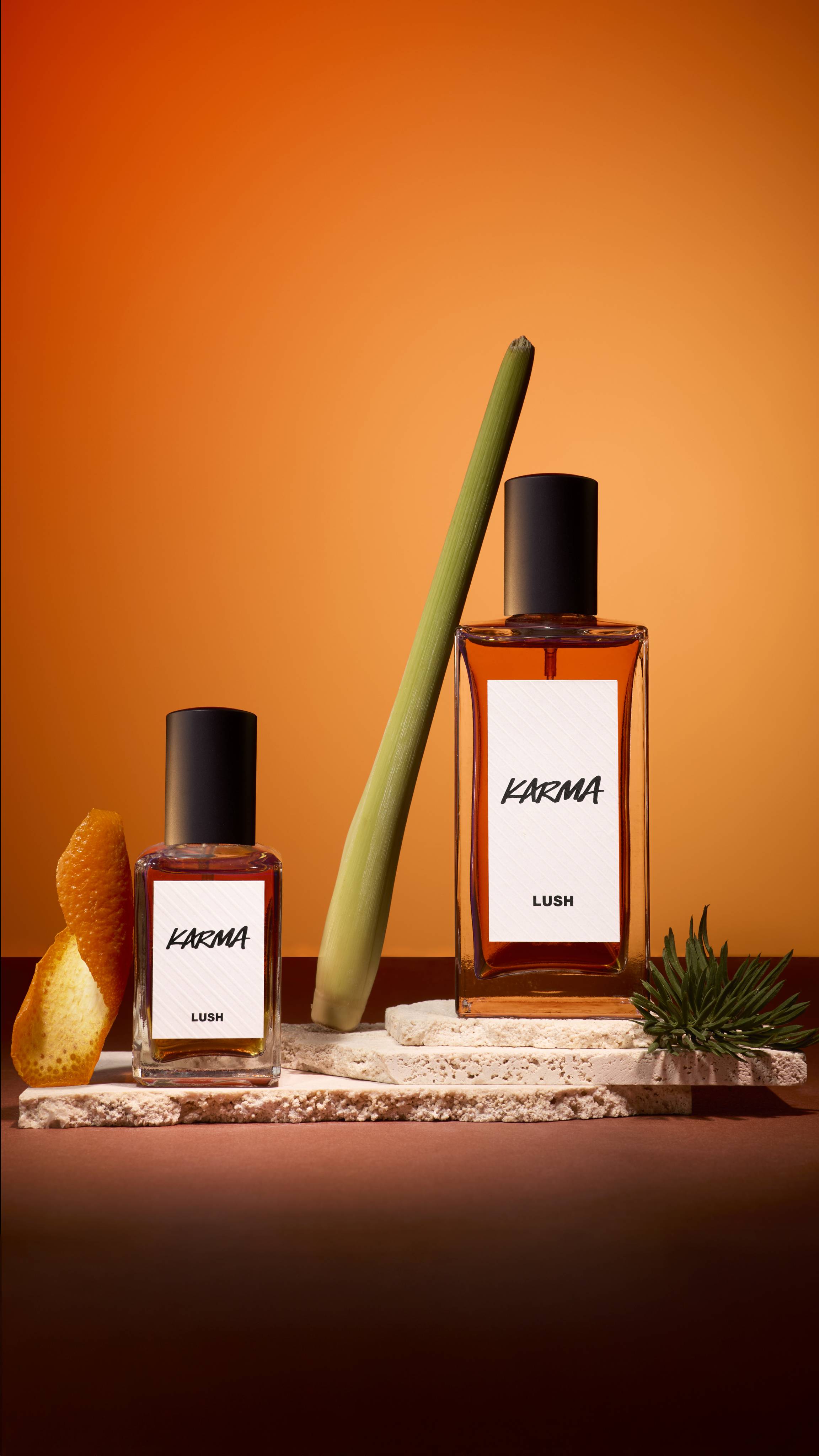Two bottles of Lush 'Karma' perfume stand on textured stone slabs against an orange background. They are styled with a curl of orange peel, a green stalk of lemongrass, and a small sprig of pine, reflecting the scent's ingredients.
