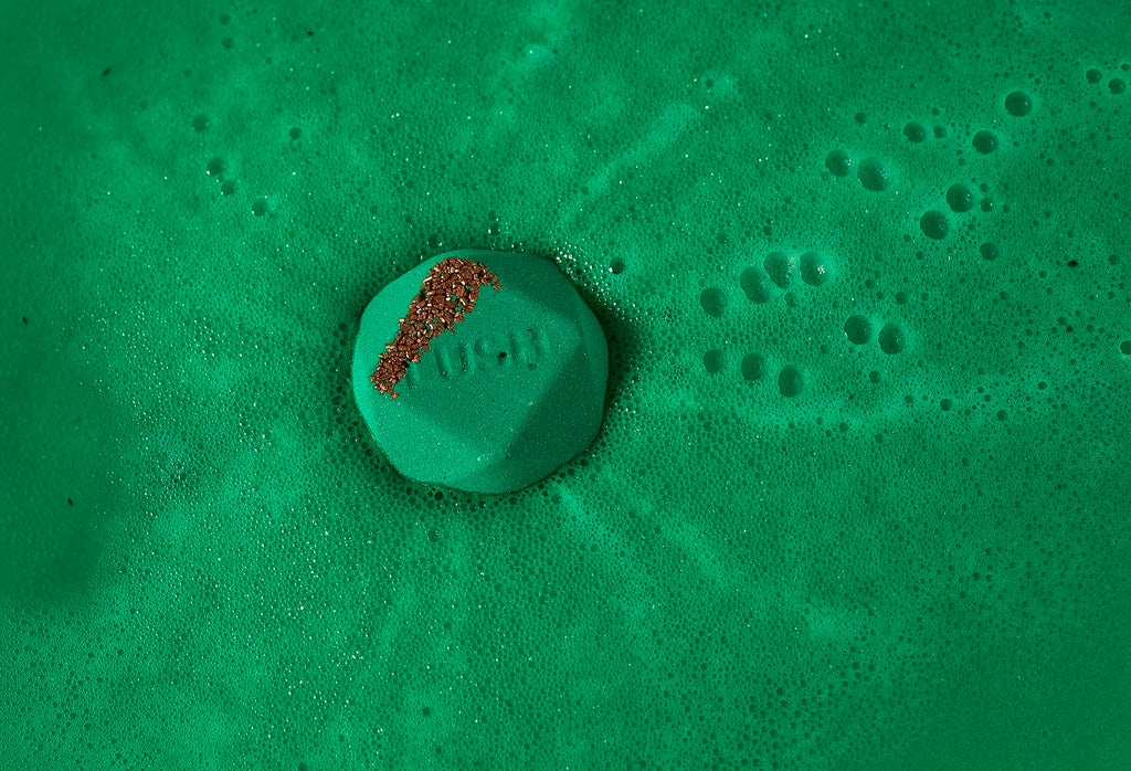 Kokage bath bomb floats in deep, emerald green sea of foam with orange-coloured salt and "Lush" embossed on top.