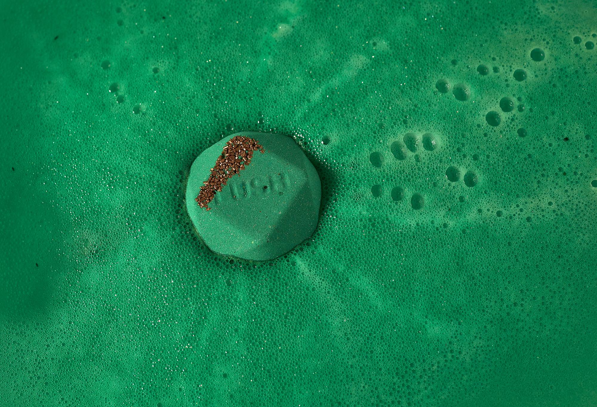 Kokage bath bomb floats in deep, emerald green sea of foam with orange-coloured salt and "Lush" embossed on top.