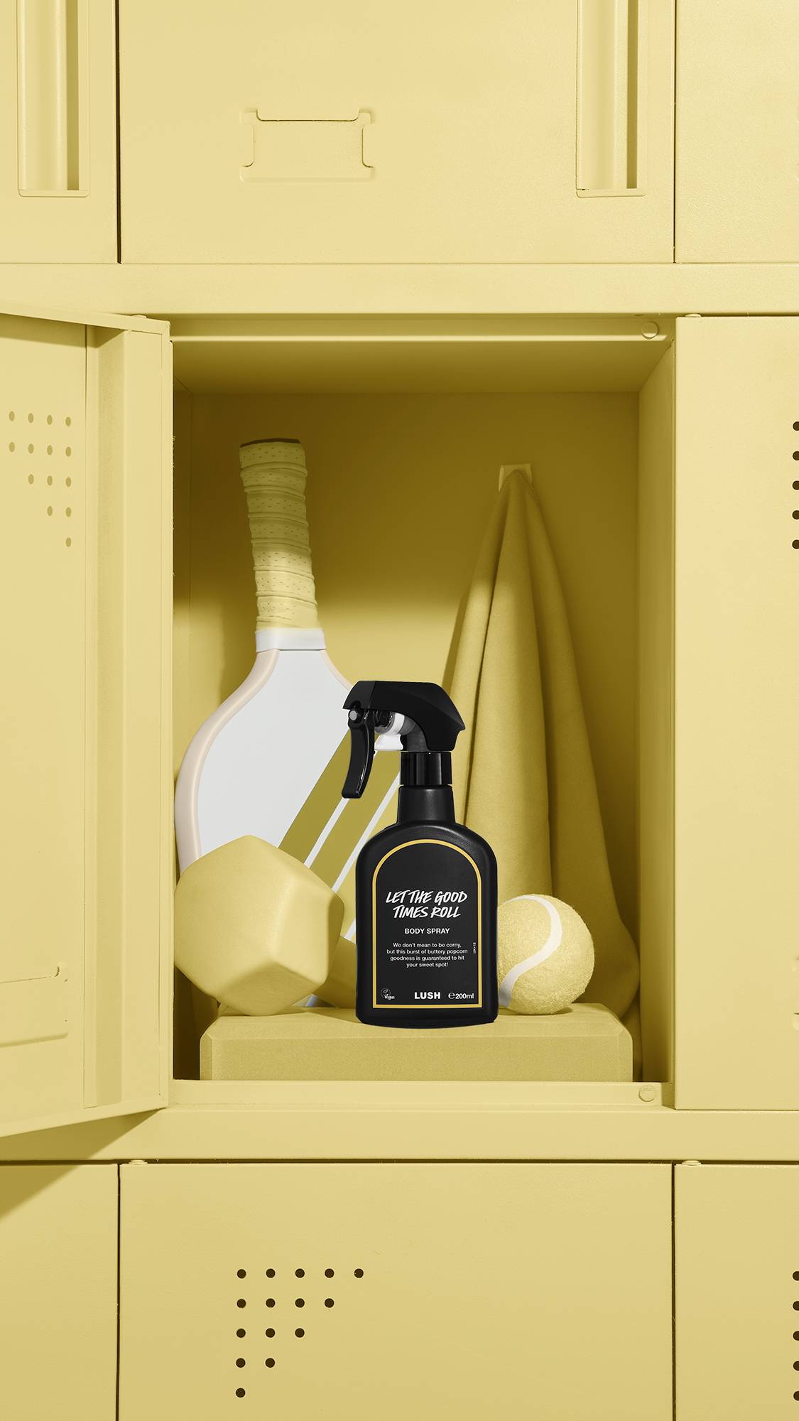 A black Lush body spray bottle labelled 'Let The Good Times Roll' sits inside an open yellow locker. The locker also contains a sports racket, a yellow tennis ball, a yellow geometric shape, and a hanging towel.