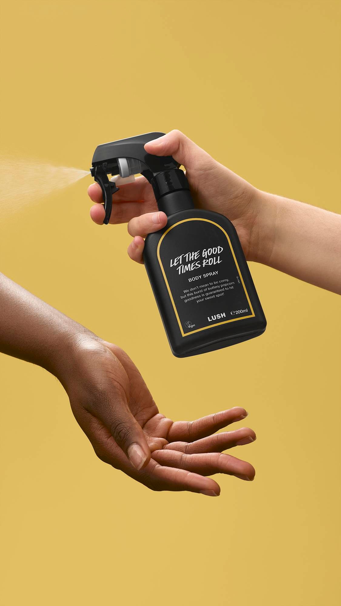 Against a solid yellow background, a hand holds a black Lush bottle labelled 'Let The Good Times Roll', releasing a fine mist of body spray towards another person's open hand.