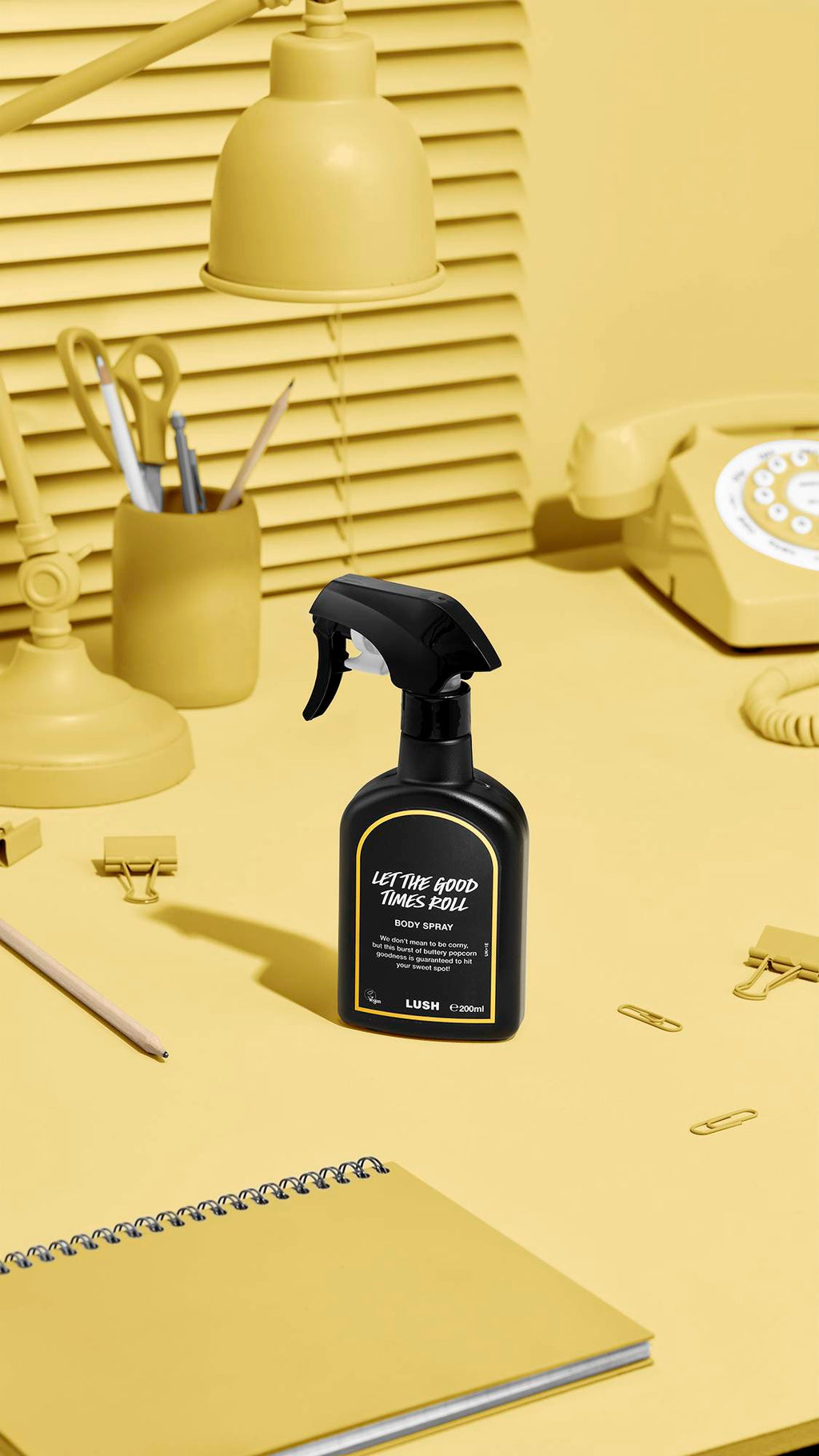A black Lush body spray bottle labelled 'Let The Good Times Roll' stands on a yellow desk. The monochromatic background features matching yellow items: a desk lamp, a pen pot, stationery, a spiral notebook, and a retro rotary telephone.