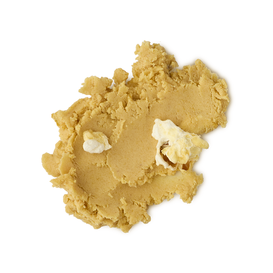 A close-up, overhead view of a thick, crumbly smudge of product (likely a scrub or balm) in a warm tan or yellow-gold color. Two small, lighter creamy white pieces are embedded in the texture, all set against a light grey background.