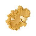 A close-up, overhead view of a thick, crumbly smudge of product (likely a scrub or balm) in a warm tan or yellow-gold color. Two small, lighter creamy white pieces are embedded in the texture, all set against a light grey background.