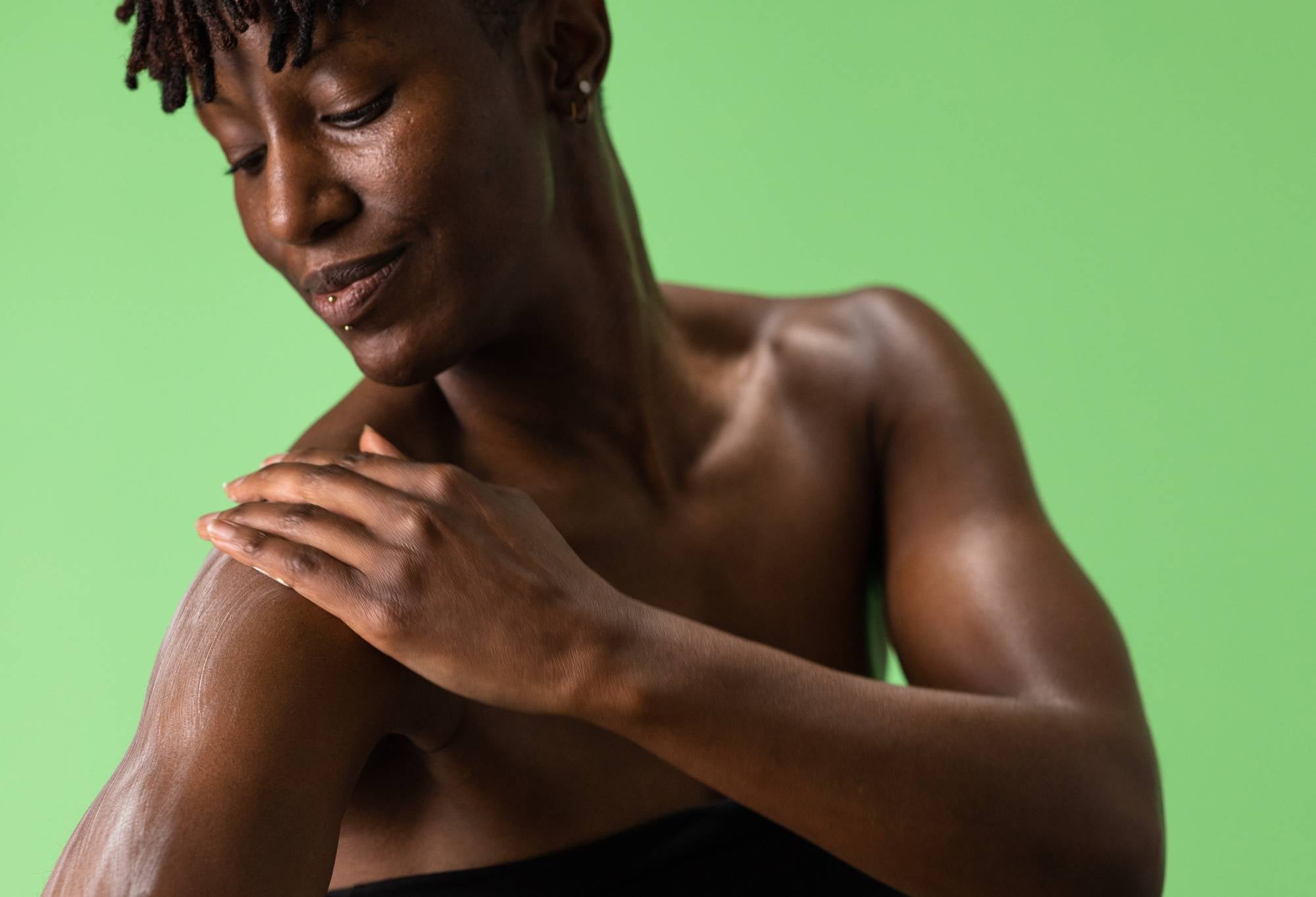 A smiling person wrapped in a towel looks down at their shoulder, as they rub a light green body butter onto their arm.