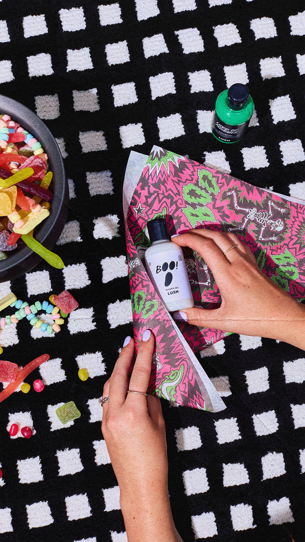 A person's hands wrap a Lush 'BOO!' shower gel bottle in vibrant magenta and green Halloween Lokta paper. The items rest on a black and white chequered rug, with a green bottle and a bowl of colourful jelly sweets nearby.