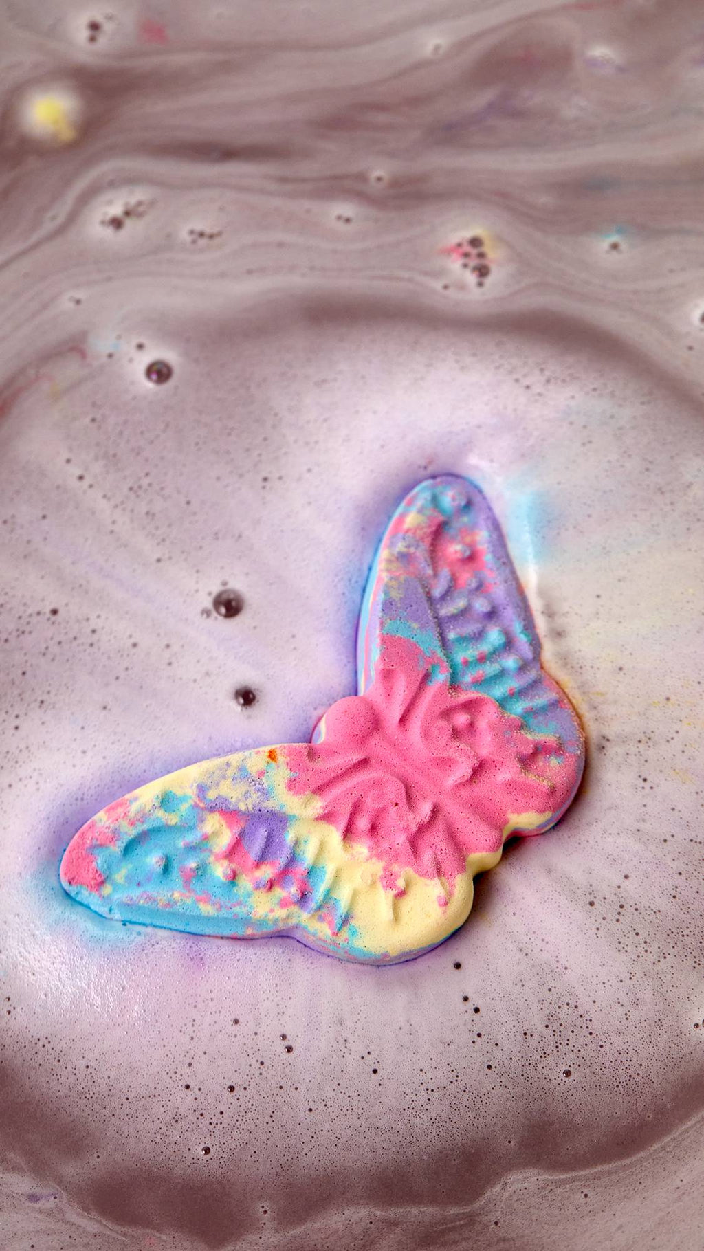 An aerial view of the colourful, Luna's Butterfly bath bomb as it dissolves in the bath. The product is giving off a layer of colourful, rippling, velvety foam across the surface of the water.