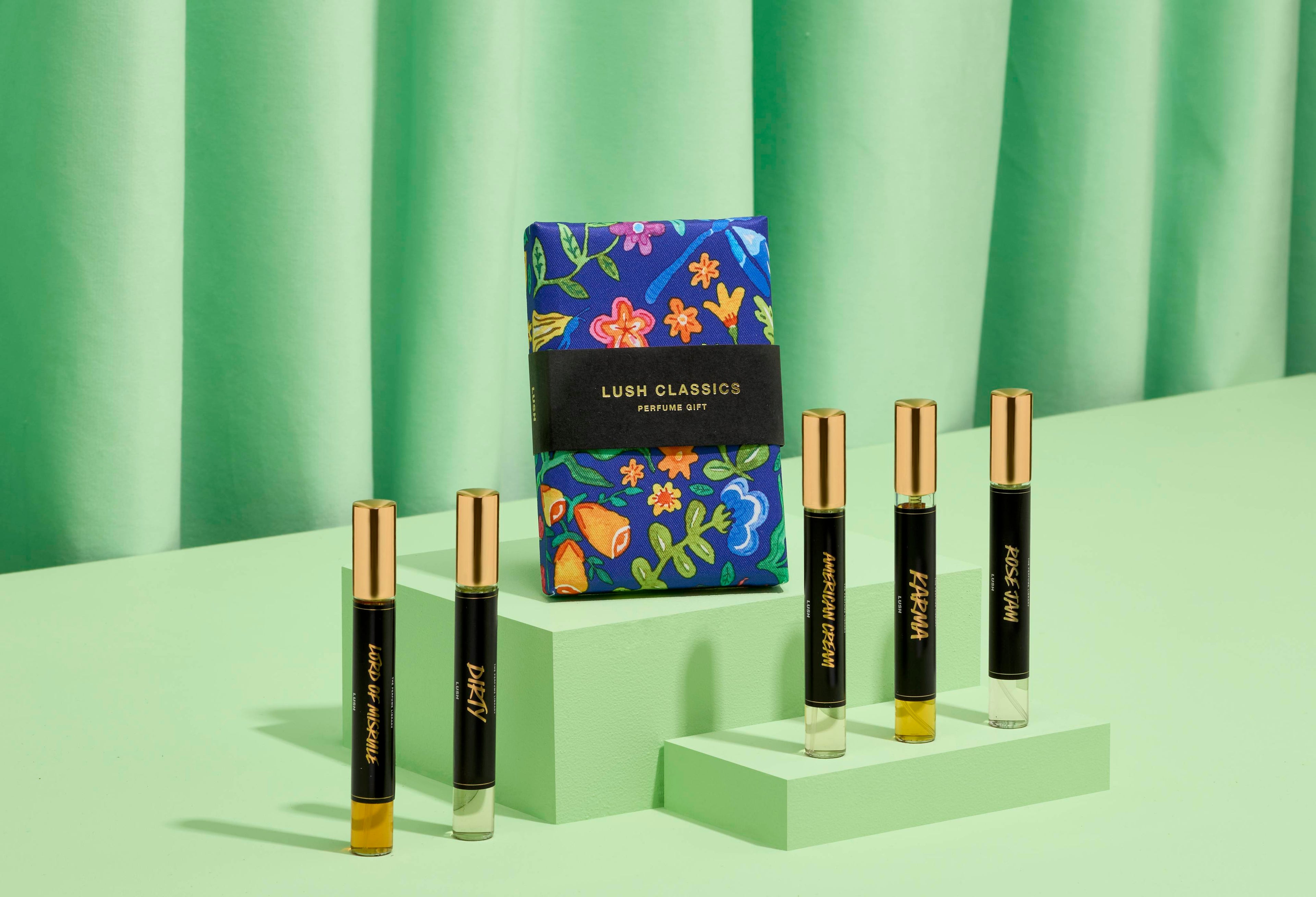 The image shows a "LUSH CLASSICS" perfume gift set: five thin, cylindrical glass bottles on light green steps. A vibrant blue box with floral designs sits behind them. Matching green fabric is draped for the background.