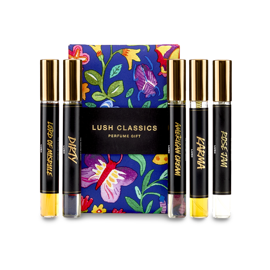 The image shows a "LUSH CLASSICS" perfume gift set. Five thin, cylindrical glass perfume bottles with amber and clear liquids inside are arranged around a dark blue box featuring vibrant floral and butterfly illustrations.