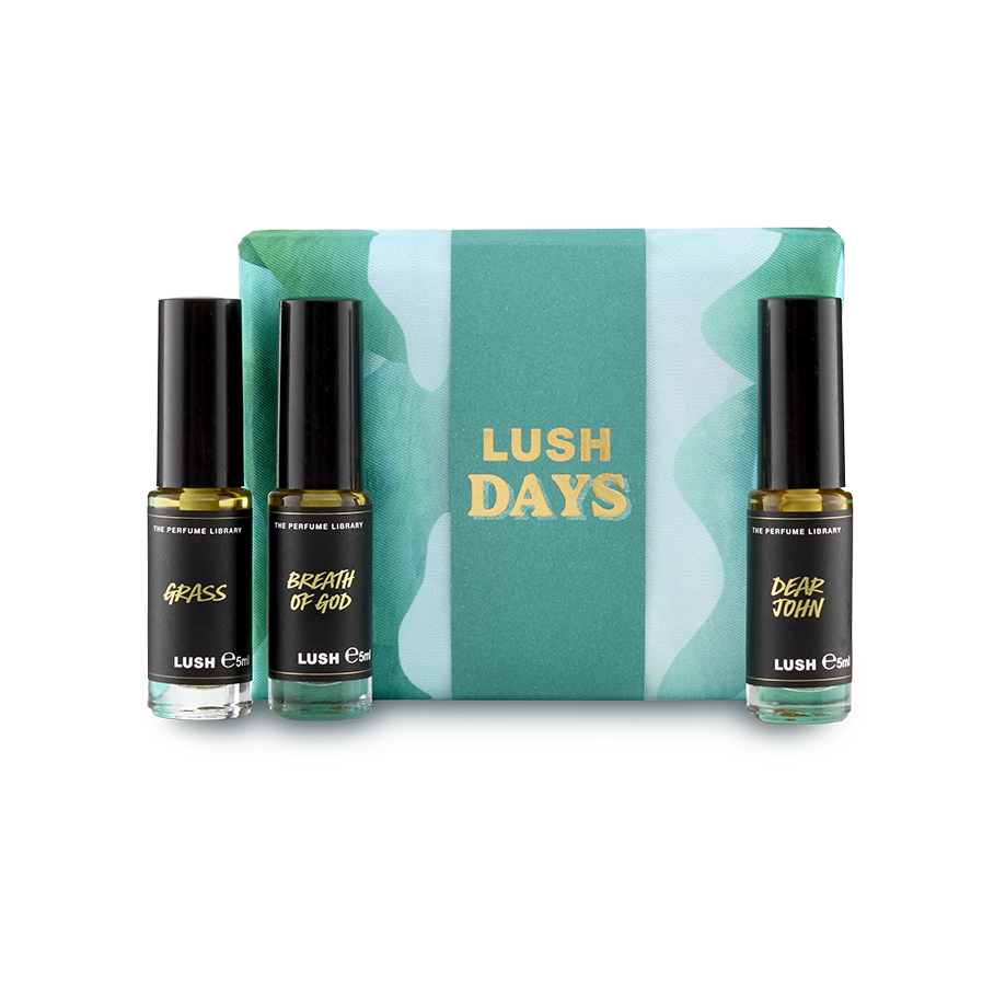 The image shows a "LUSH DAYS" perfume gift set. Three cylindrical glass perfume bottles are arranged around a vibrant teal box with a wavy pattern and yellow text. Perfumes include 'Grass', 'Breath of God' and 'Dear John'.