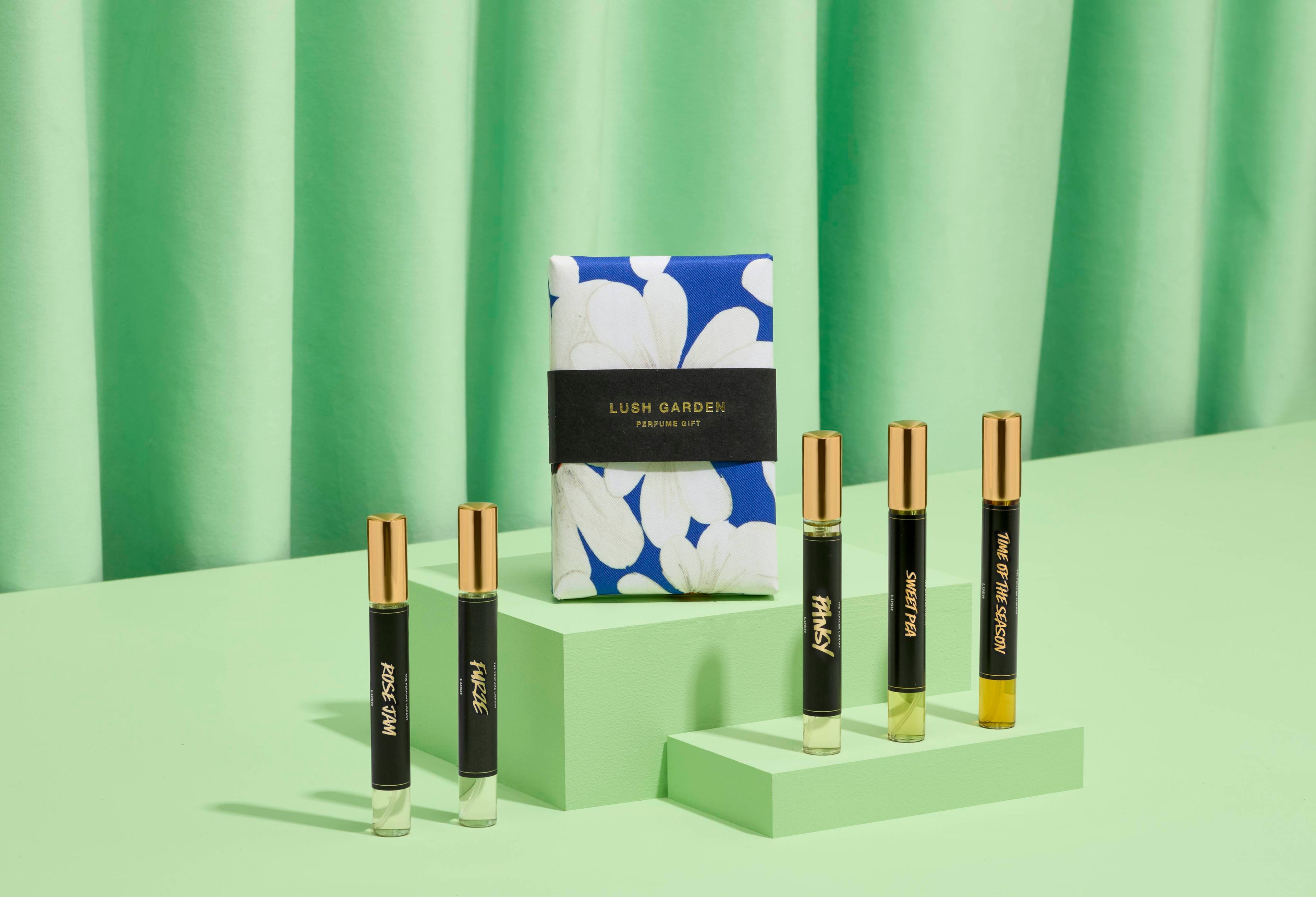 The image shows a "LUSH GARDEN" perfume gift set: five thin, cylindrical glass bottles on light green steps. A vibrant blue box with floral designs sits behind them. Matching green fabric is draped for the background.