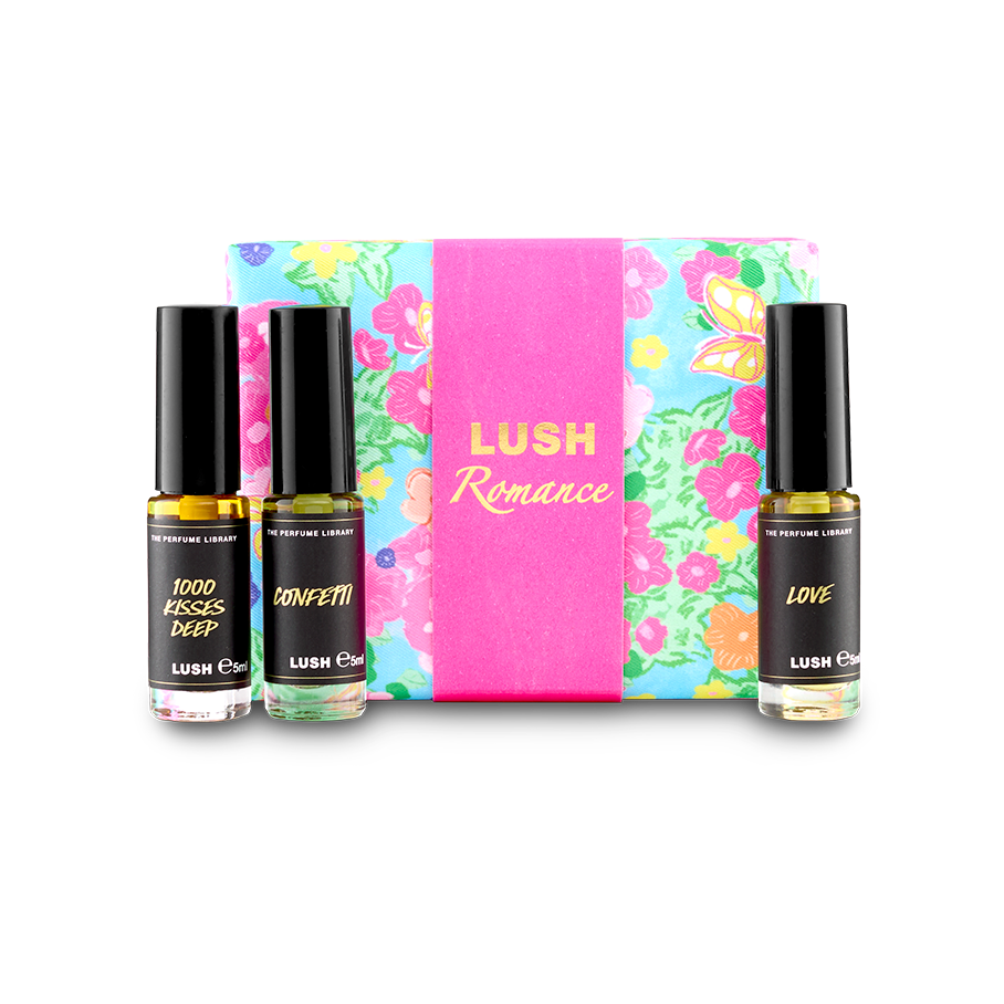 The image shows a "LUSH Romance" perfume gift set. Three thin glass perfume bottles with amber and clear liquids are arranged around a light blue box with vibrant floral patterns and a bright pink band.