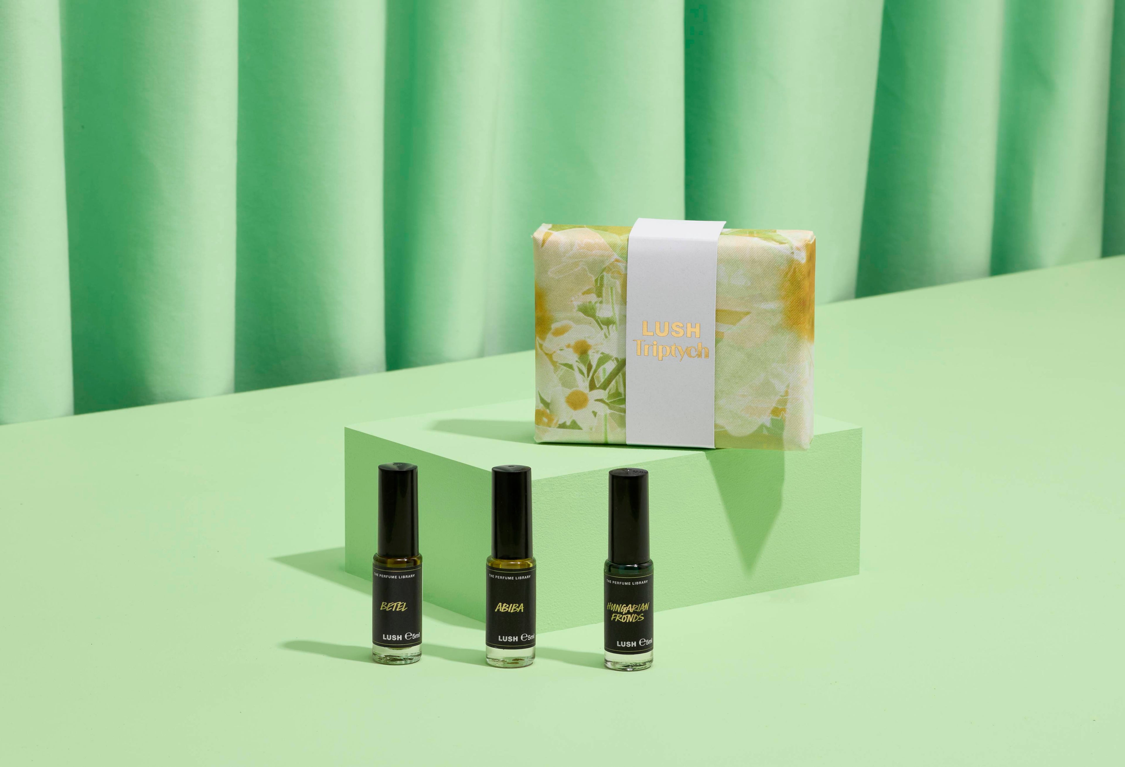 The image shows a "LUSH Triptych" perfume gift set: three thin glass bottles on light green steps. A pale yellow and green marbled box with a white band sits behind them. Matching green fabric forms the backdrop.