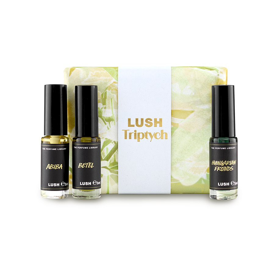 The image shows a "LUSH Triptych" perfume gift set. Three thin, cylindrical glass perfume bottles with amber and clear liquids are arranged around a pale yellow and green marbled box with a white band.