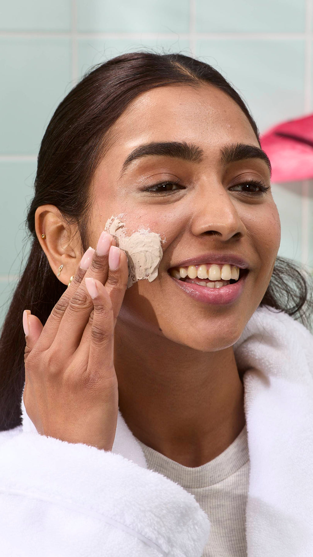 A model in a white fluffy dressing gown smiles as they apply the thick, beige-coloured "Me Time" face mask to their cheek. A pale green tiled wall is visible in the background.