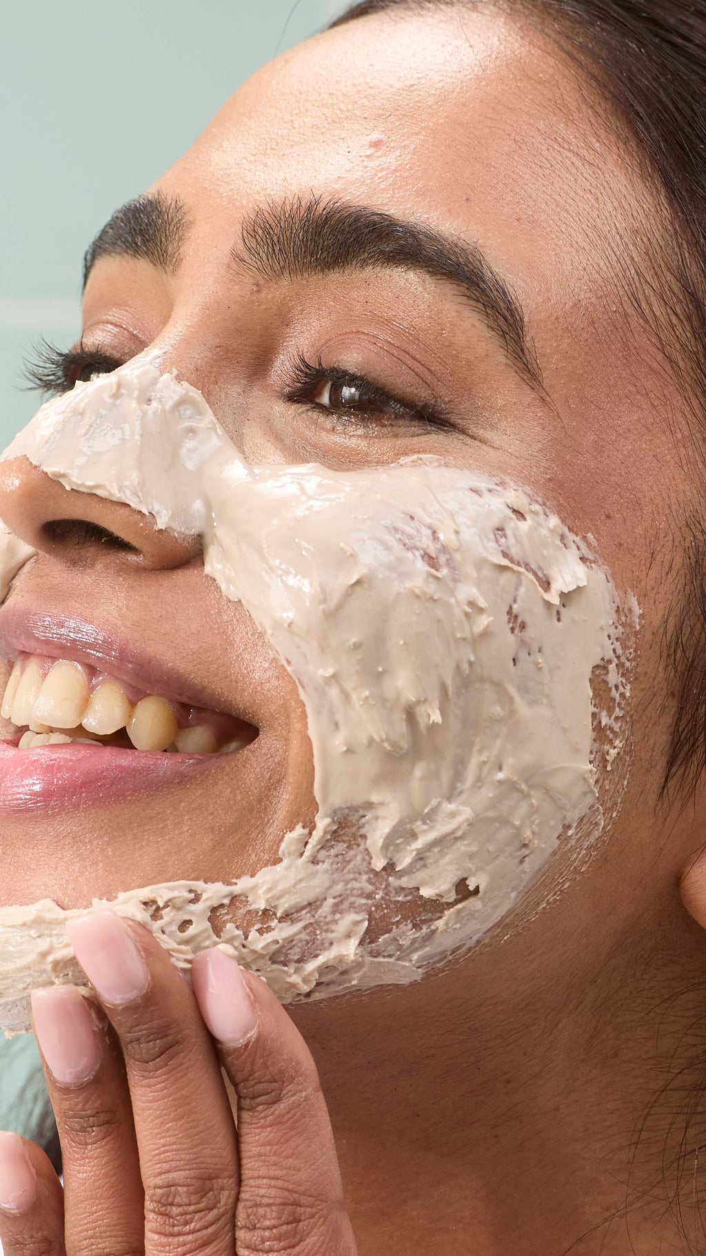 A close-up of a model smiling as they apply the thick, beige-coloured "Me Time" face mask to their cheeks and jawline. The product is spread across their skin, showing its smooth yet dense texture. Pale green tiles are in the background.