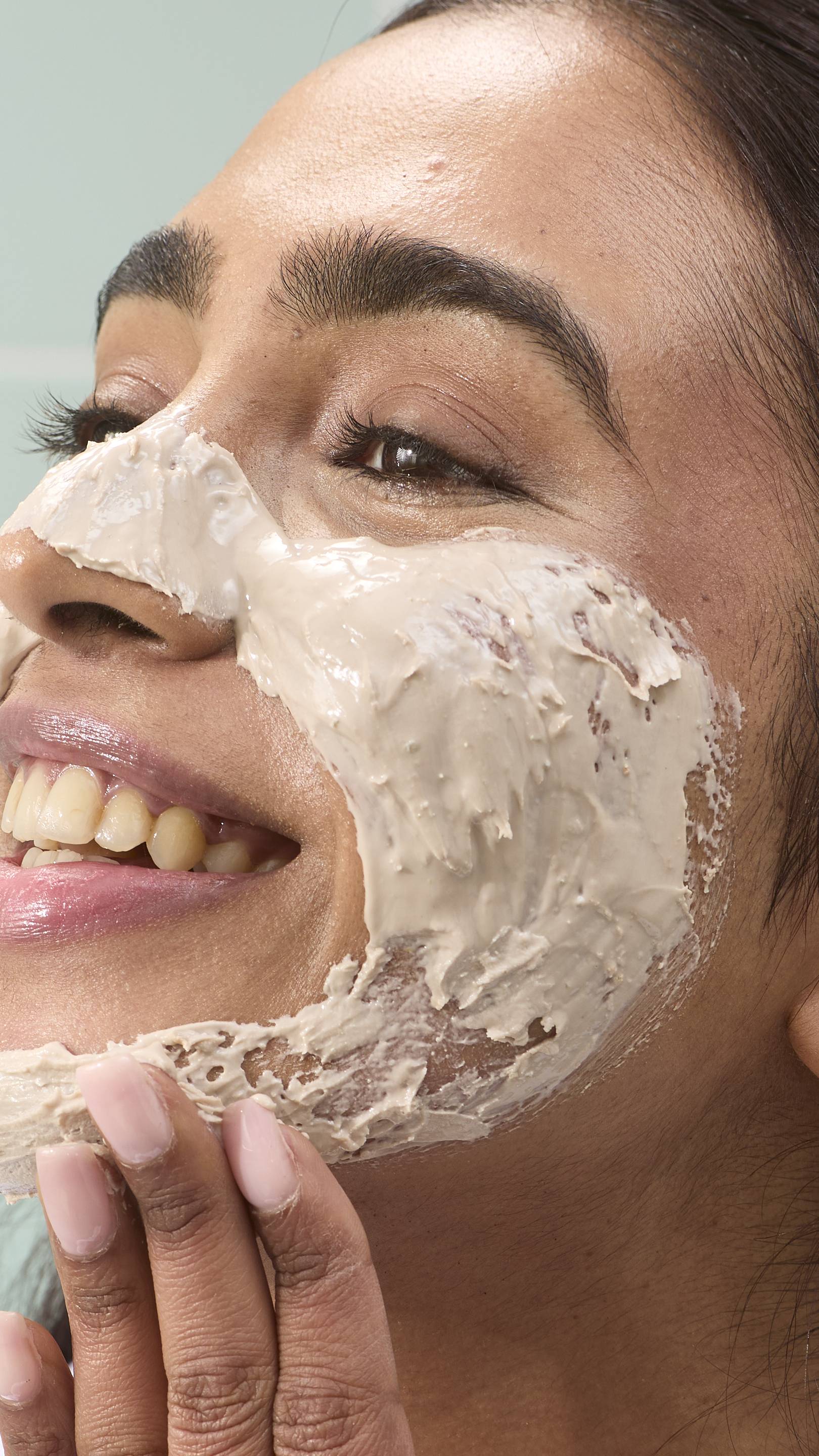 A close-up of a model smiling as they apply the thick, beige-coloured "Me Time" face mask to their cheeks and jawline. The product is spread across their skin, showing its smooth yet dense texture. Pale green tiles are in the background.